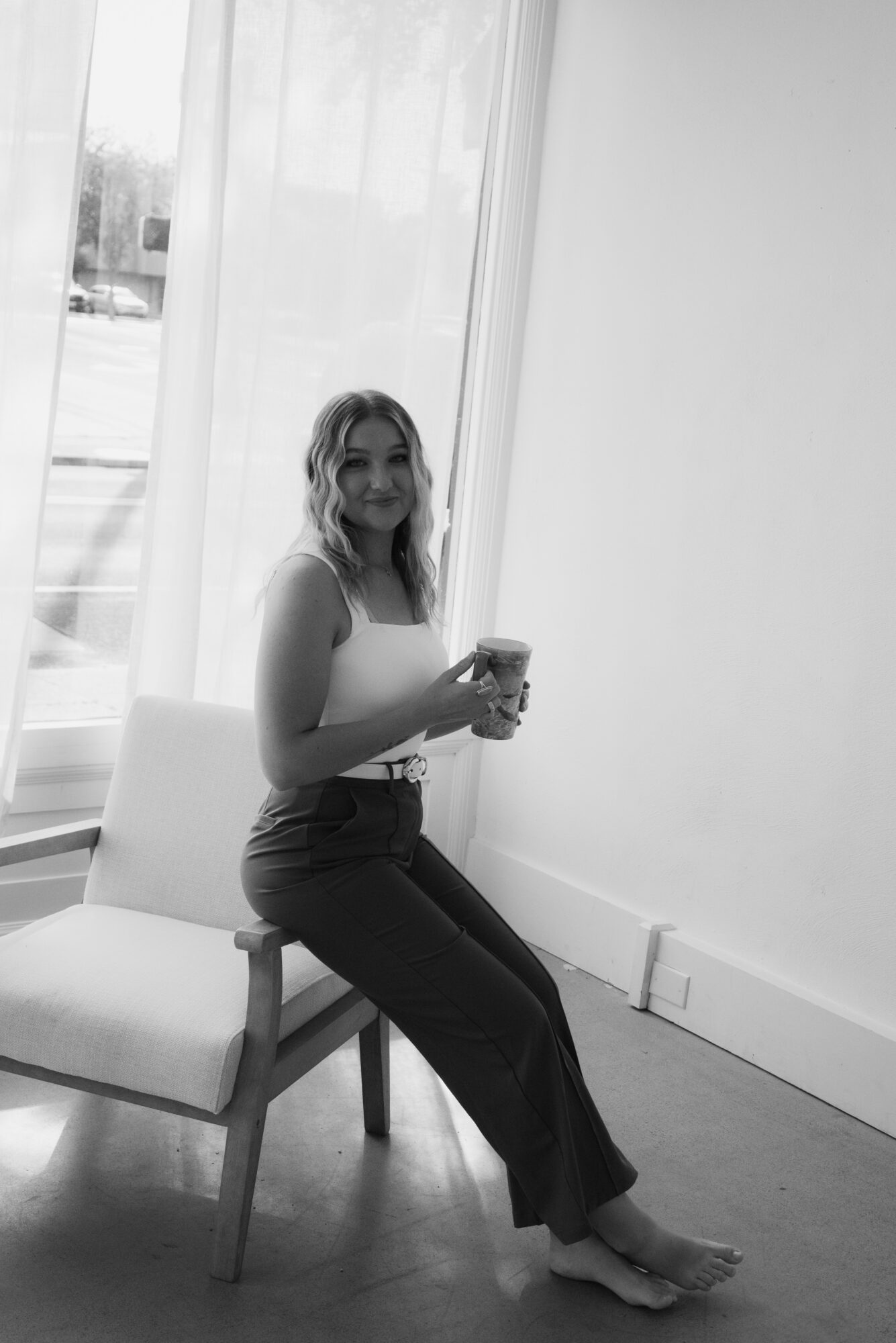Woman sitting on a chair holding a drink, smiling, near a window with curtains, in a bright room.