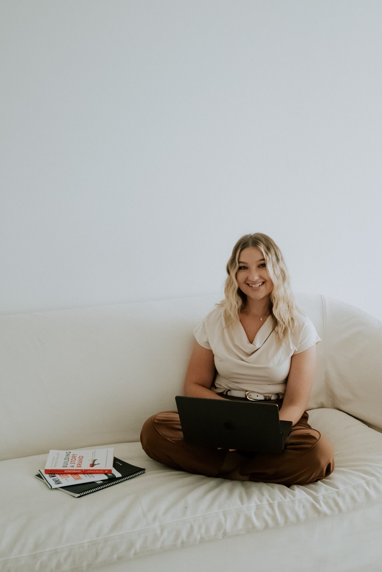 Woman sitting on a white couch with a laptop, smiling, with magazines nearby, against a plain light background.