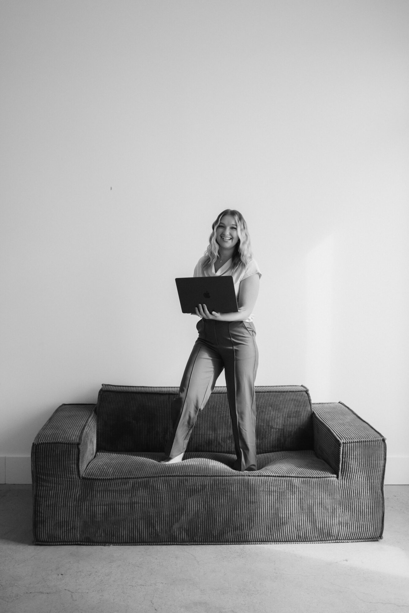 Woman standing on sofa holding a laptop, against a plain wall, in black and white.