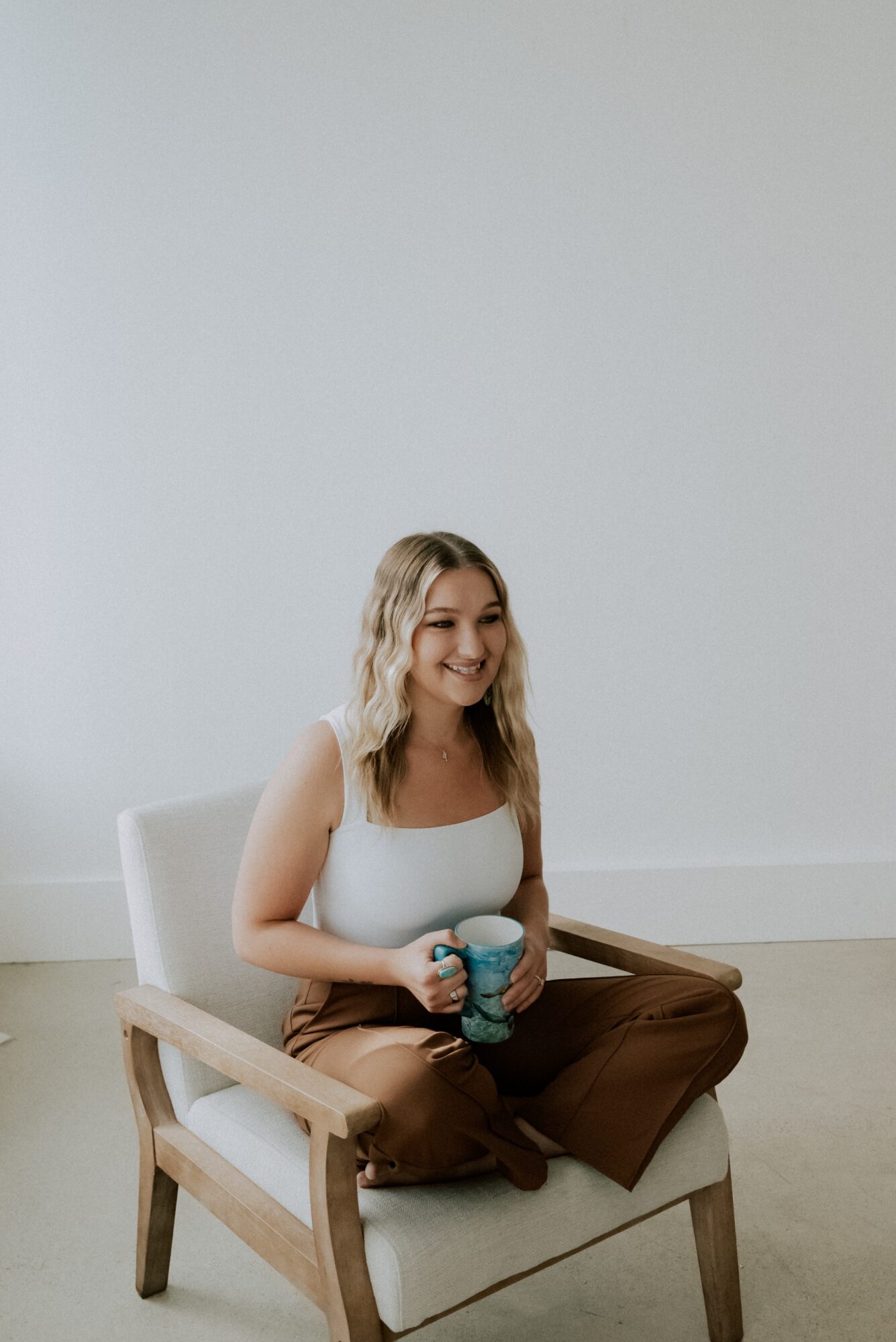 Young woman with wavy blonde hair sitting on a white chair, holding a blue mug, smiling, against a plain white wall.