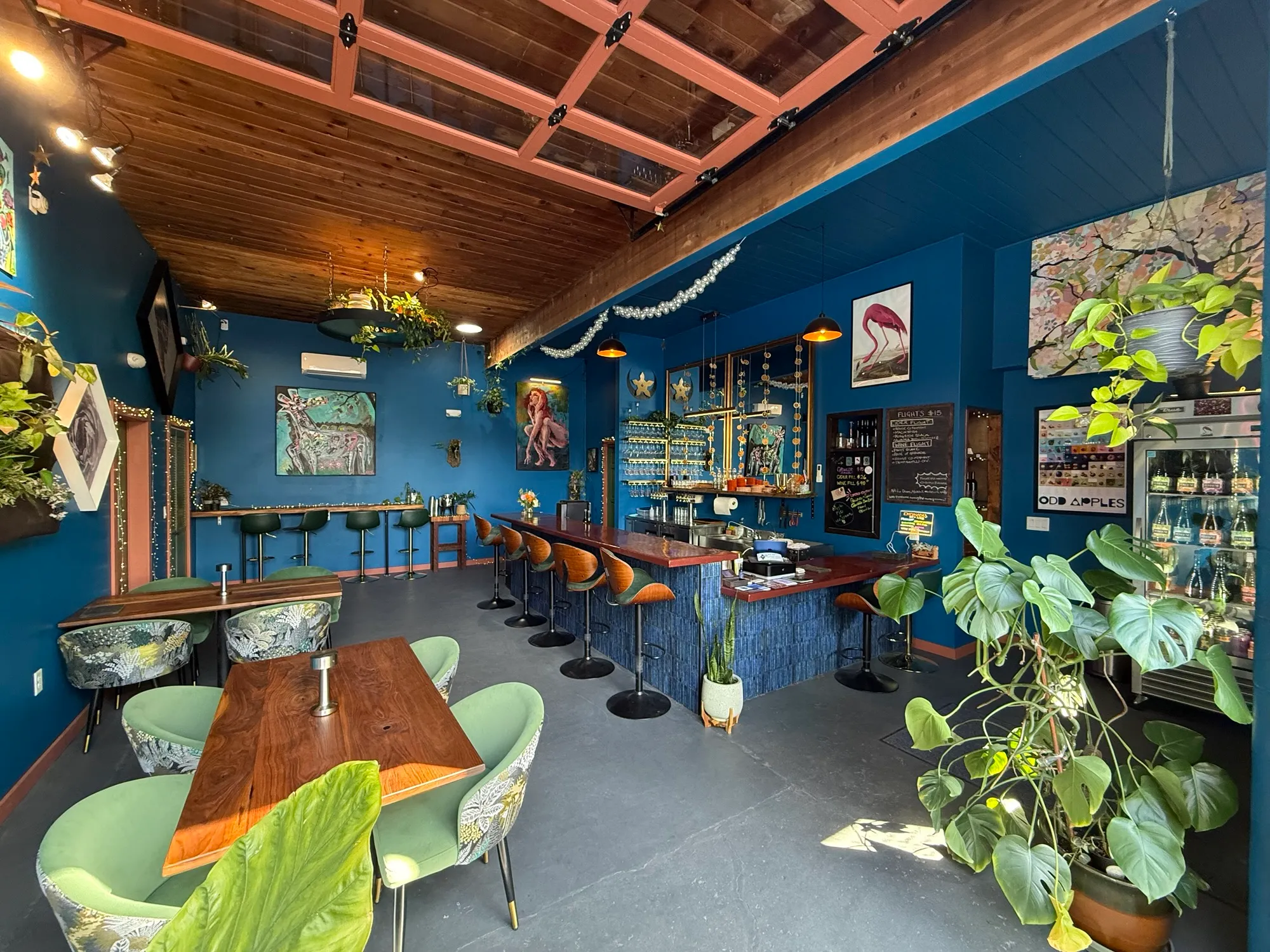 Interior of a cafe with blue walls, wooden ceiling, bar counter, seating area, and large plants near windows.