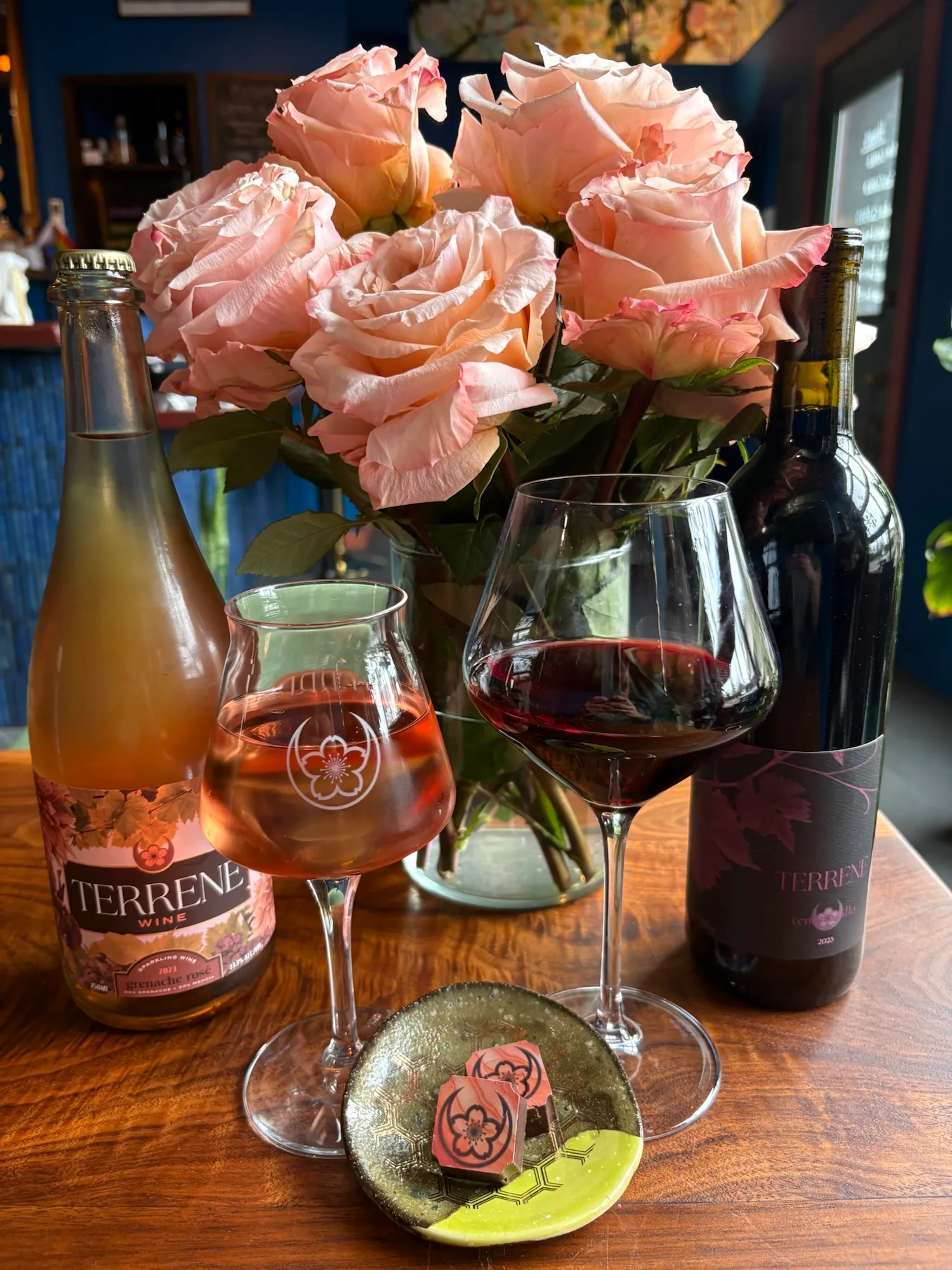 Pink roses in a vase, two wine glasses with red and rosé wine, and two bottles on a wooden table.