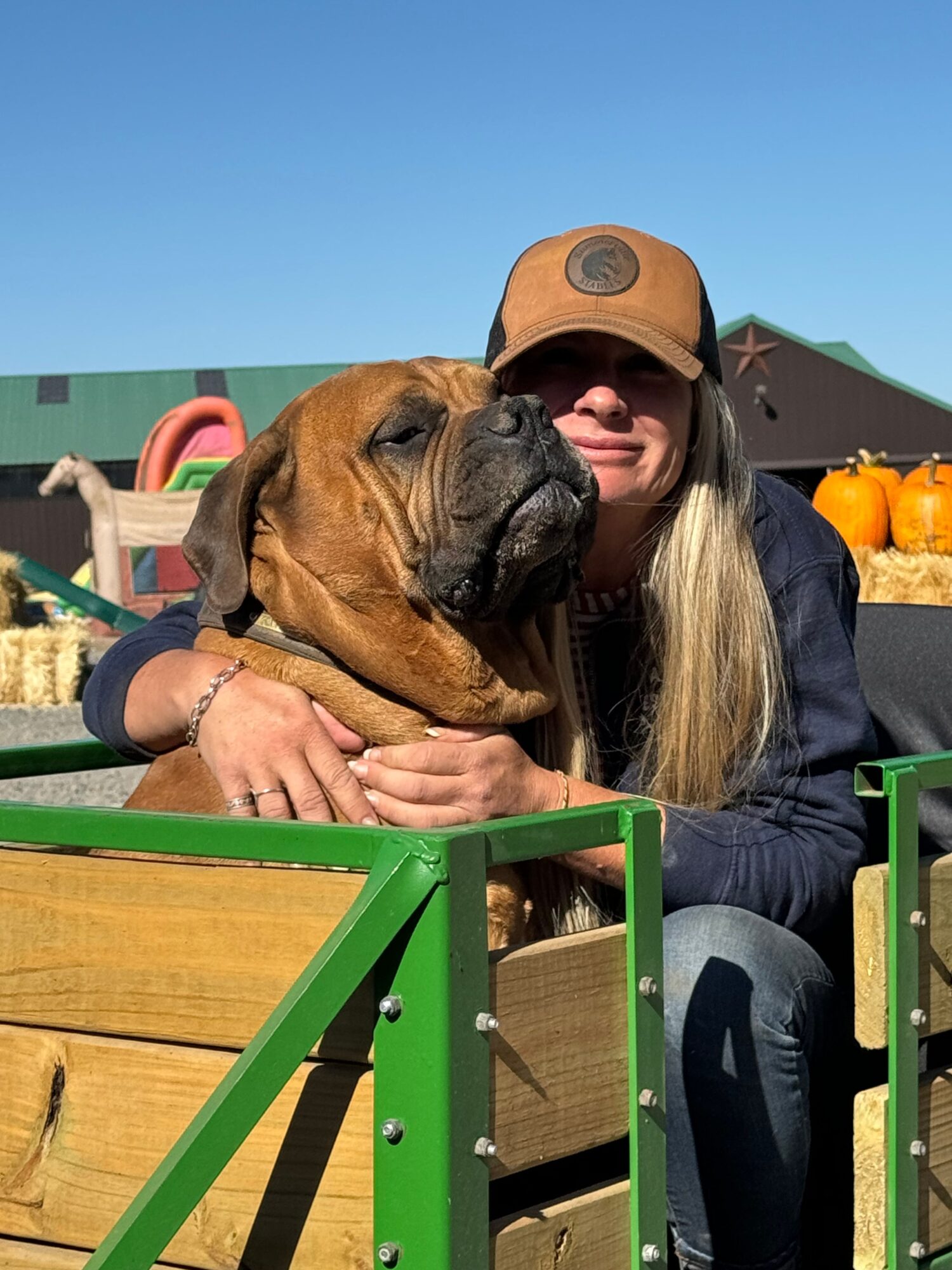 Person with long blonde hair wearing a brown cap hugging a brown dog outdoors, with a green structure nearby.