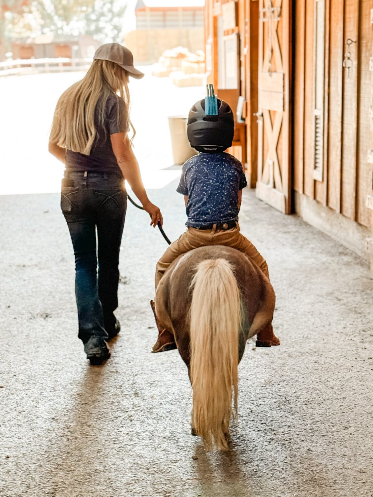 A woman walks beside a child riding a small horse inside a stable, with wooden walls and a bright background.