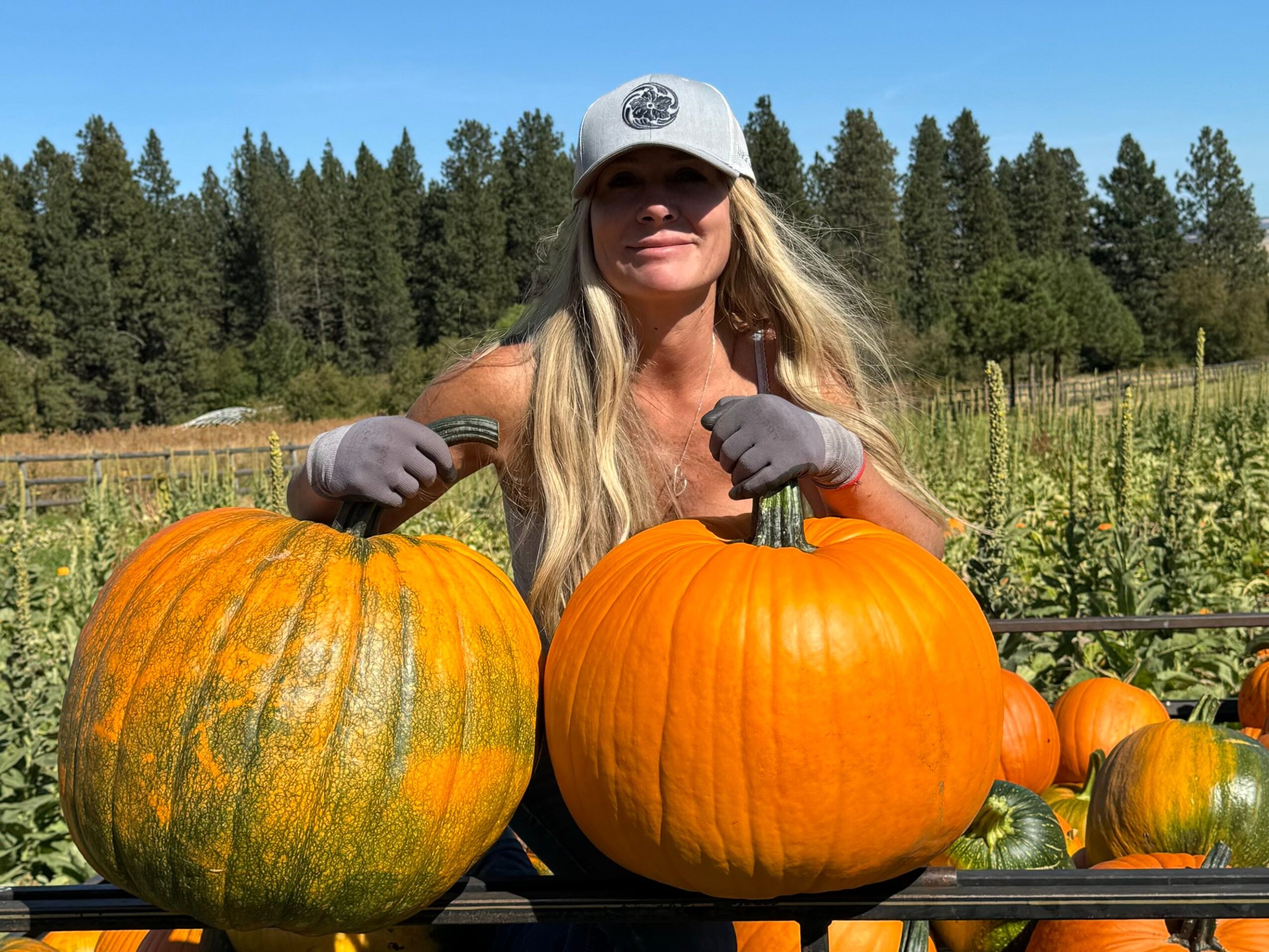 Woman with long blonde hair wearing a white cap and gray gloves, standing outdoors with pumpkins, trees, and blue sky.