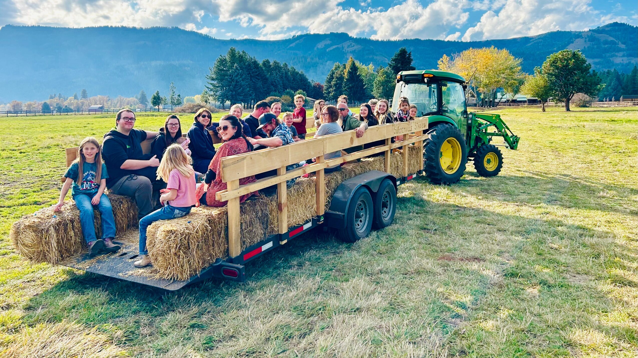 Group of children and adults sitting on a hayride trailer attached to a tractor in a field with mountains in the background.