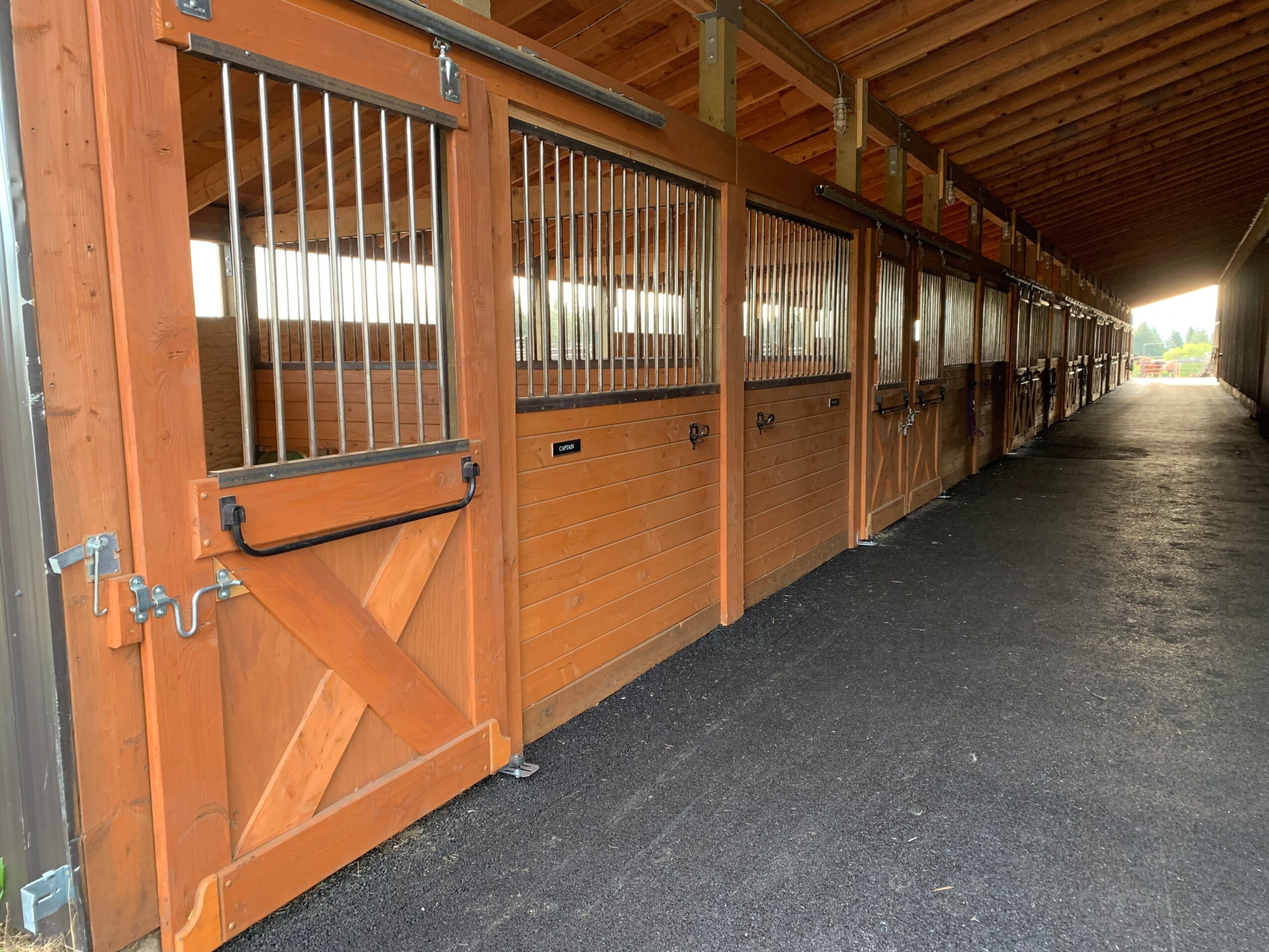 Row of wooden horse stalls with metal bars, under a wooden roof, on a paved surface, extending into the distance.