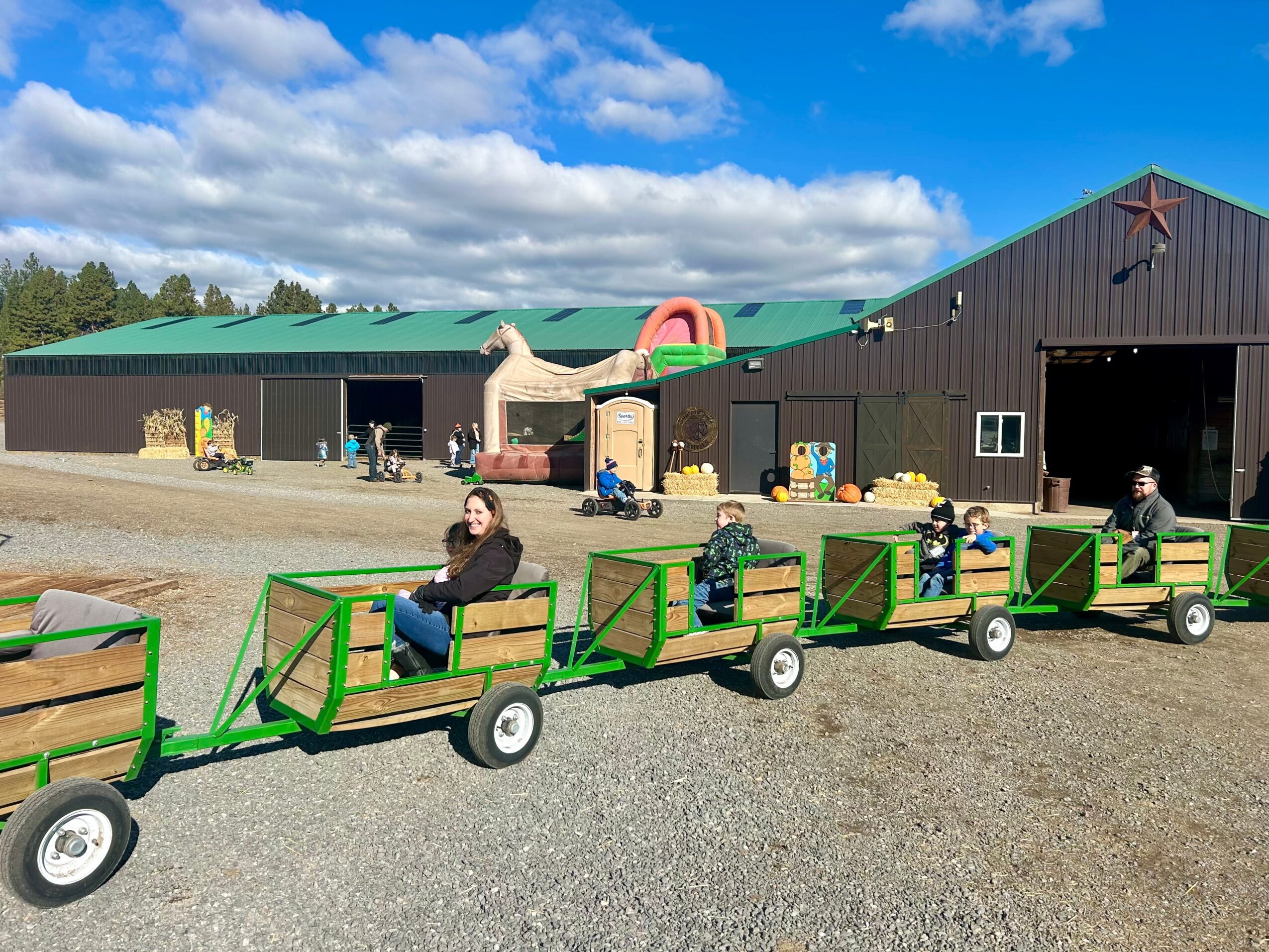 Children and adults ride a small green train outside a barn with a horse sculpture and playground equipment, under a partly cloudy sky.
