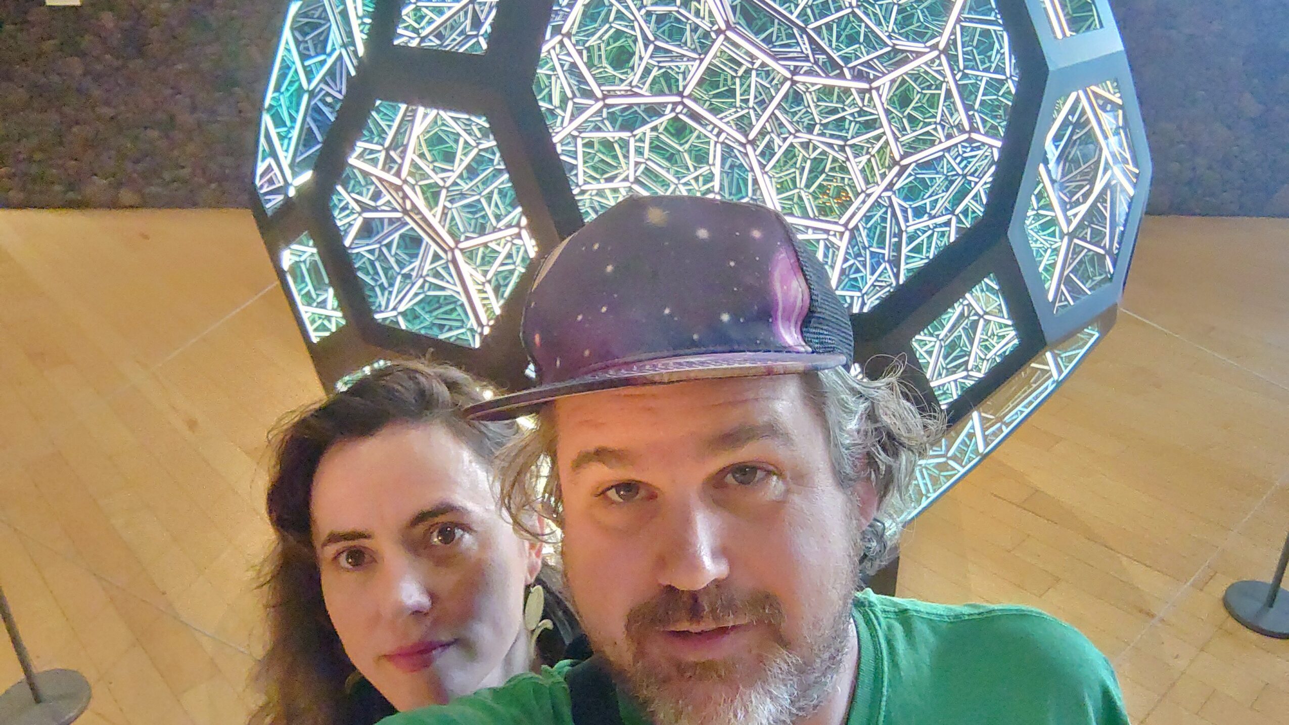 Two people taking a selfie in front of a geometric, illuminated art installation, with a wooden floor visible.