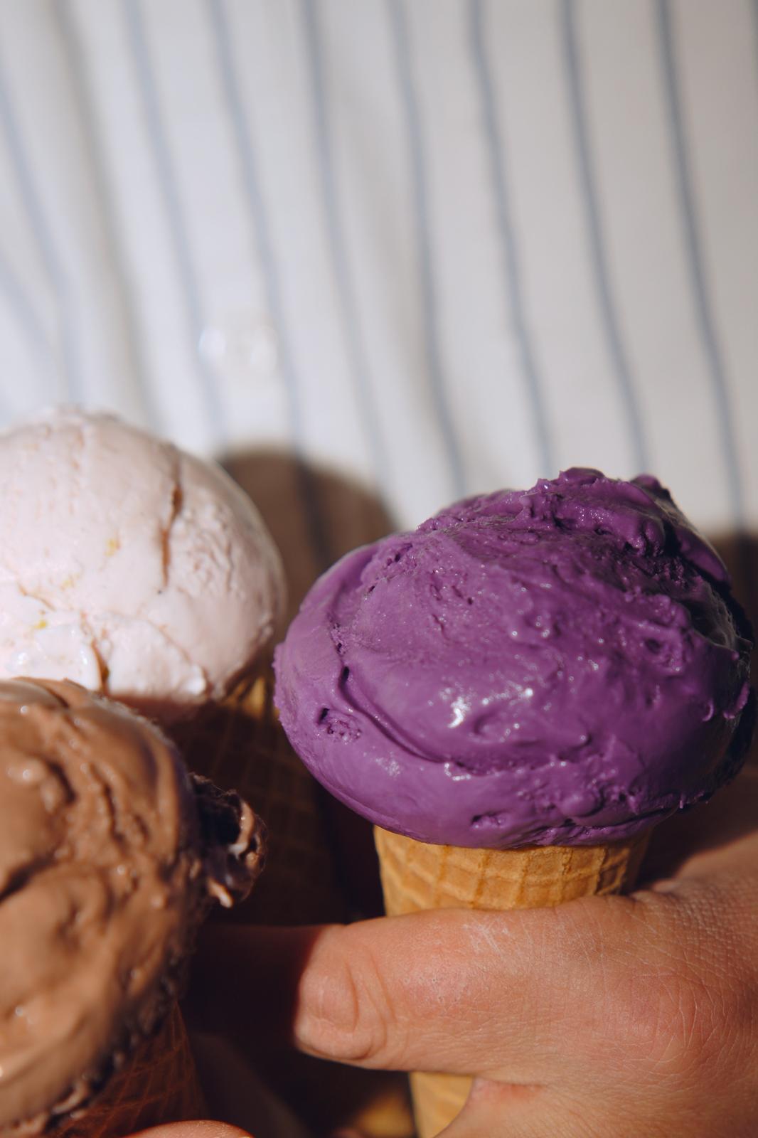 Hand holding a purple ice cream cone with other ice creams in the background, against a striped wall.