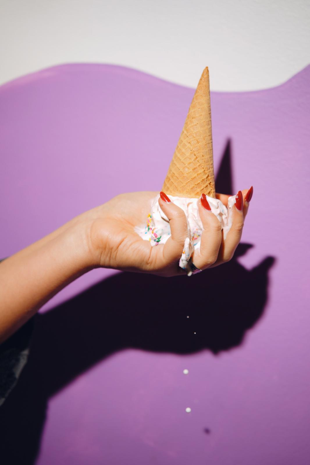 Hand holding an ice cream cone against a purple background, shadow cast on wall, with painted nails and rings.
