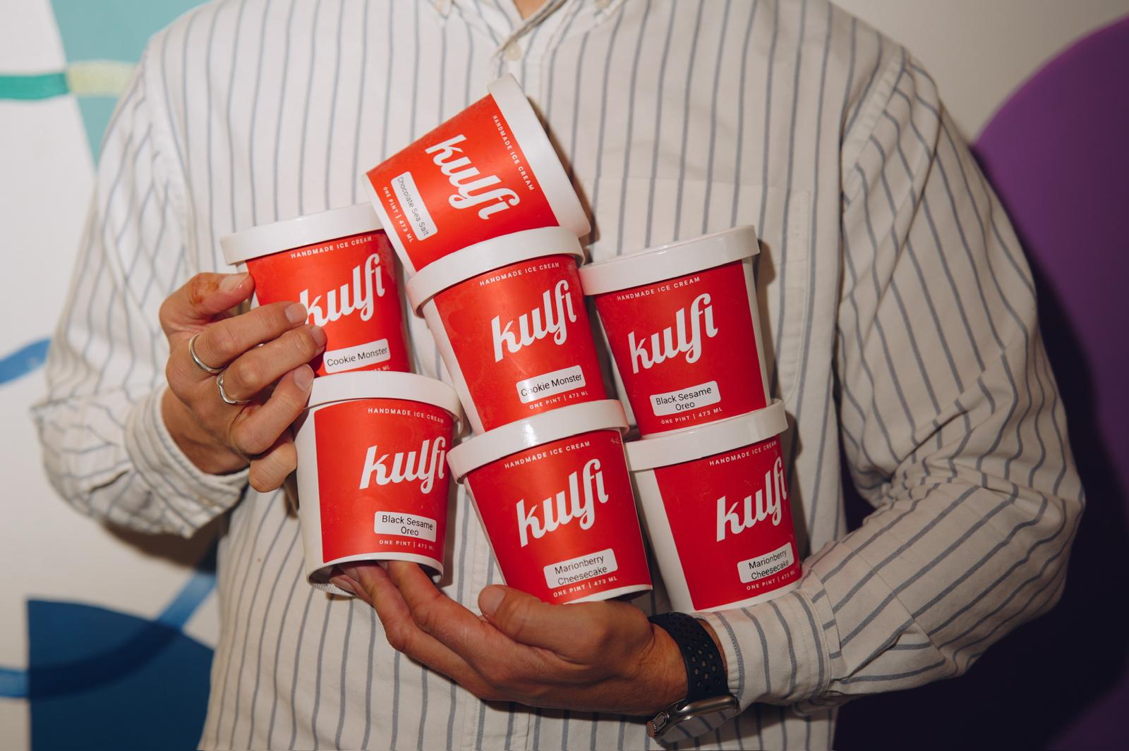 Person holding six red and white containers labeled 'kulf' in front of a colorful background.
