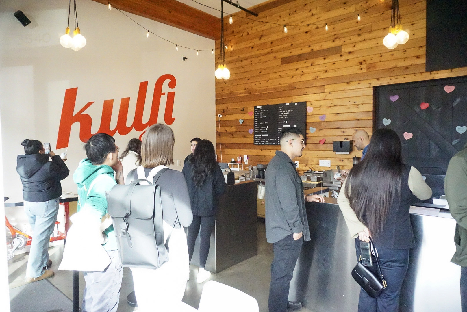 People line up at a counter inside a cafe with wooden walls and hanging lights, with a large red 'kulfi' sign on the wall.
