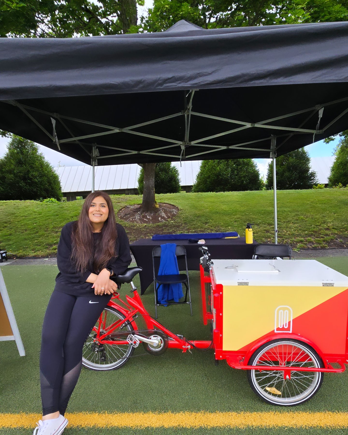 Woman standing next to a red and white food cart under a black canopy in a park, smiling, with trees and grass in background.