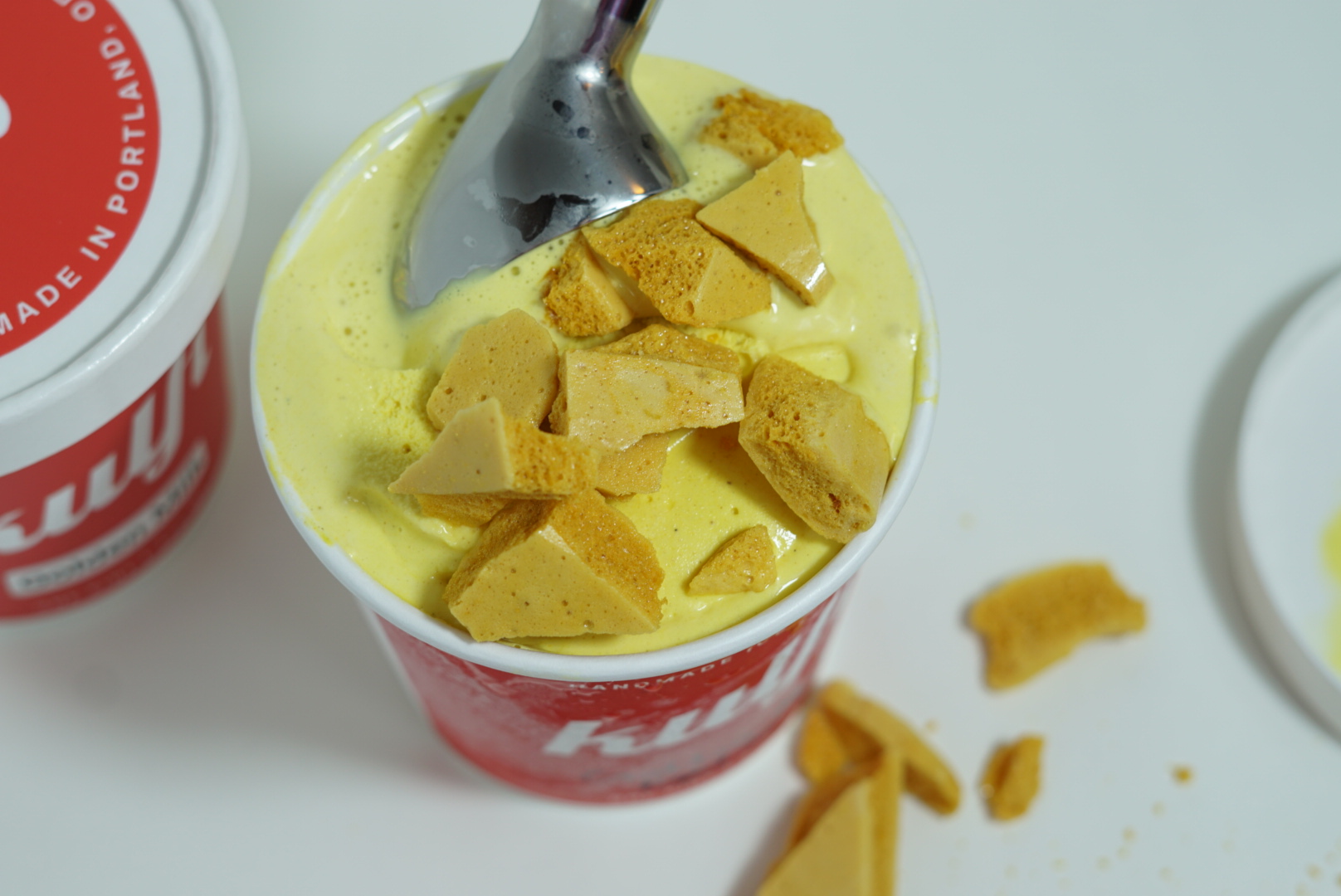 Cup of yellow ice cream topped with yellow cookie pieces, with a spoon inserted, on a white surface.