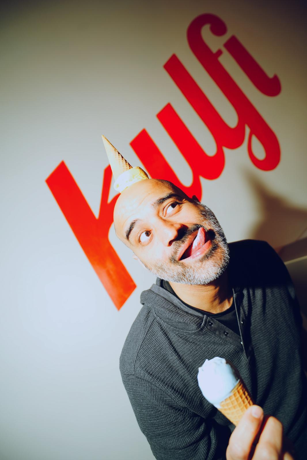 Man with a unicorn horn headband holding an ice cream cone, smiling, in front of a wall with red text.