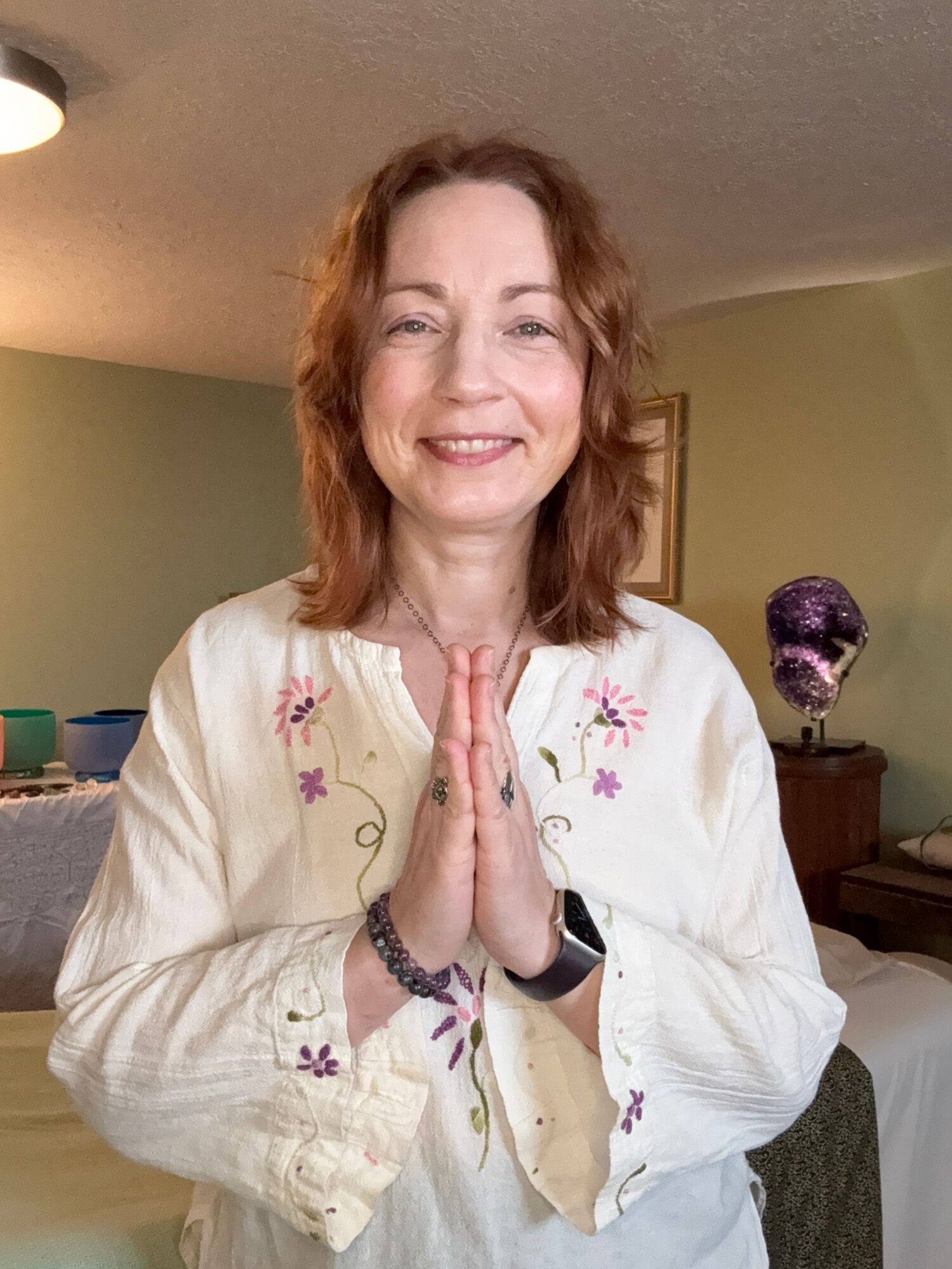 Woman with shoulder-length wavy hair smiling, wearing a white embroidered blouse, sitting indoors with a bed and purple vase in background.