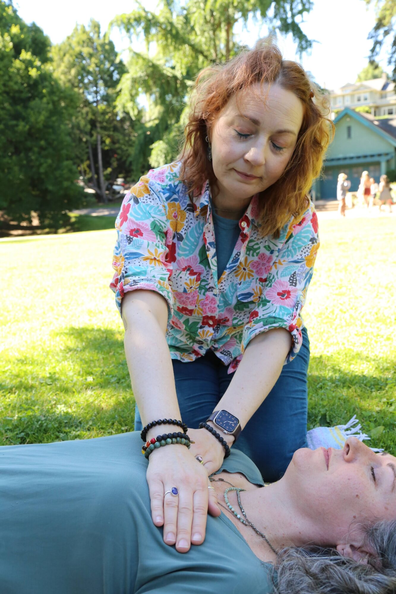 Two women outdoors, one with curly red hair and colorful blouse, the other with gray hair and blue shirt, engaging in a healing or meditative activity.