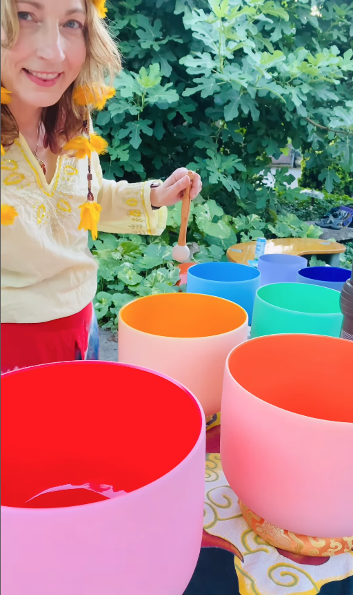 Person with a flower necklace standing outdoors with colorful singing bowls on a table, surrounded by green foliage.