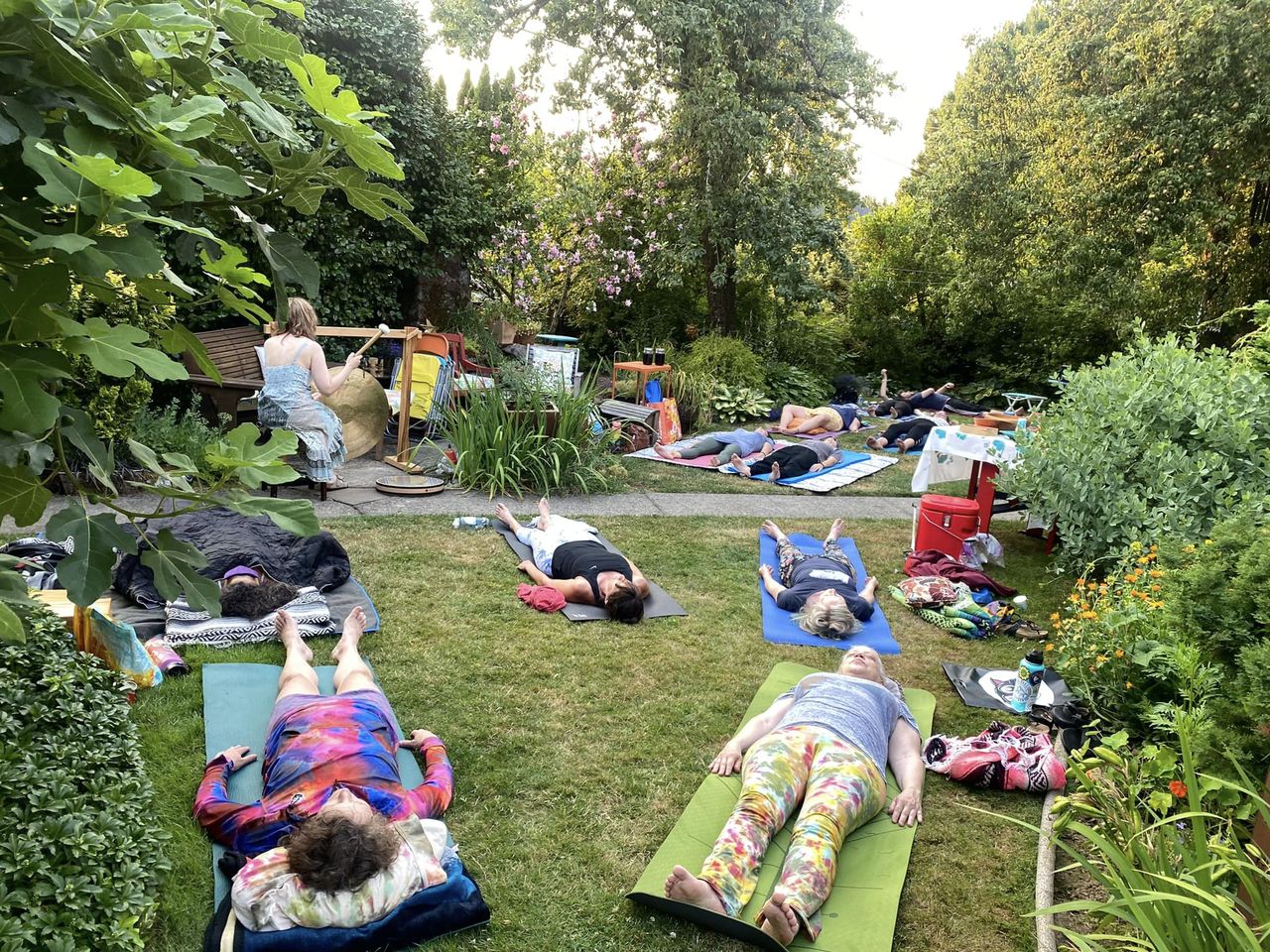 People relaxing on mats and towels in a lush garden with trees and bushes, some lying down and others sitting.