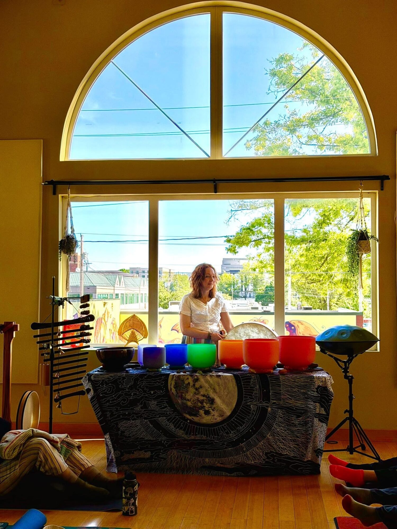 Person standing behind a table with colorful bowls, large window with trees outside, yellow walls, and sunlight.