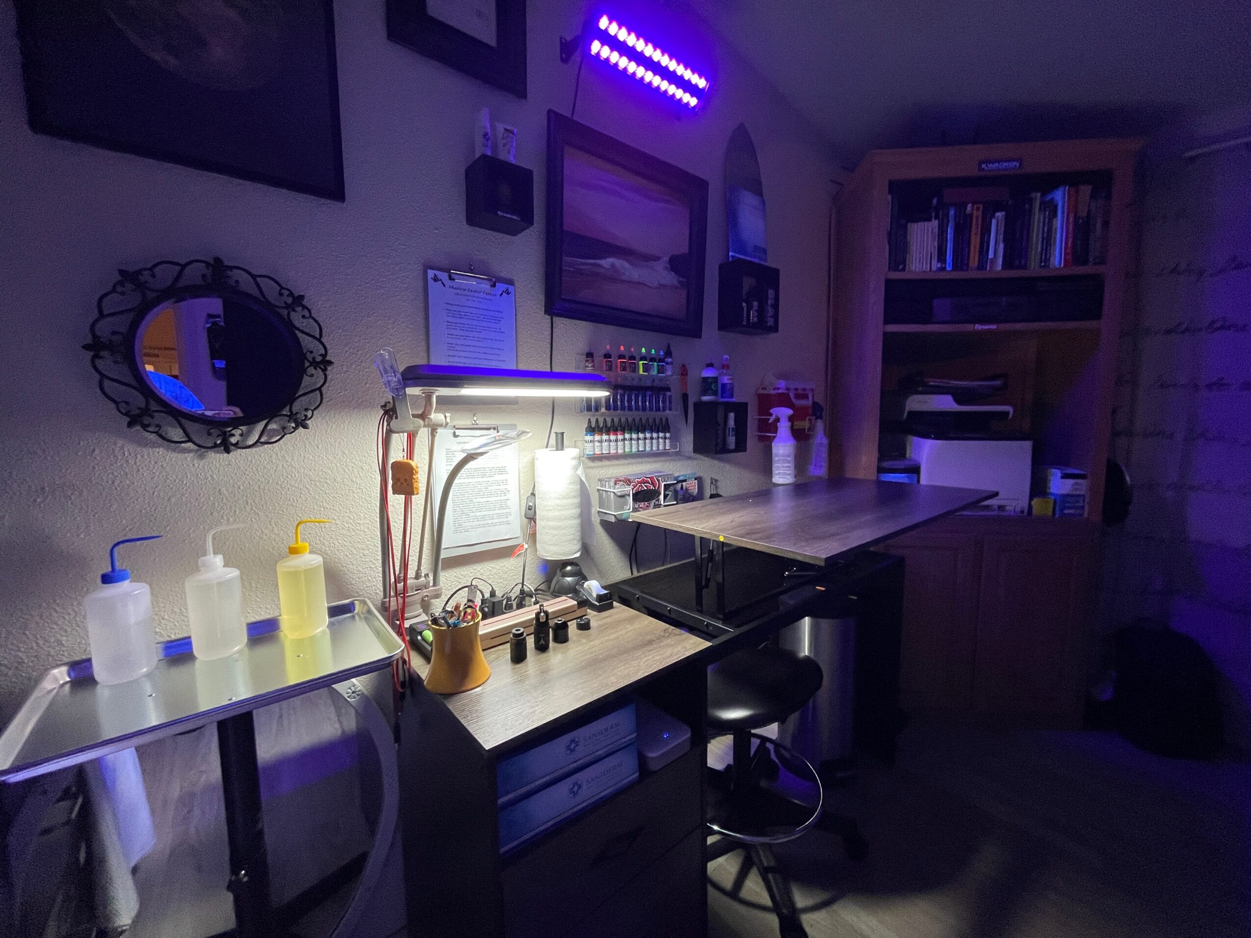 A room with a work desk, shelves, and various bottles, illuminated by purple and white lights, with a mirror on the wall.