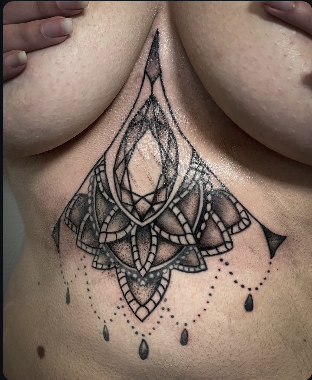 Tattoo of a geometric lotus flower with hanging teardrop shapes on the chest, between the breasts.