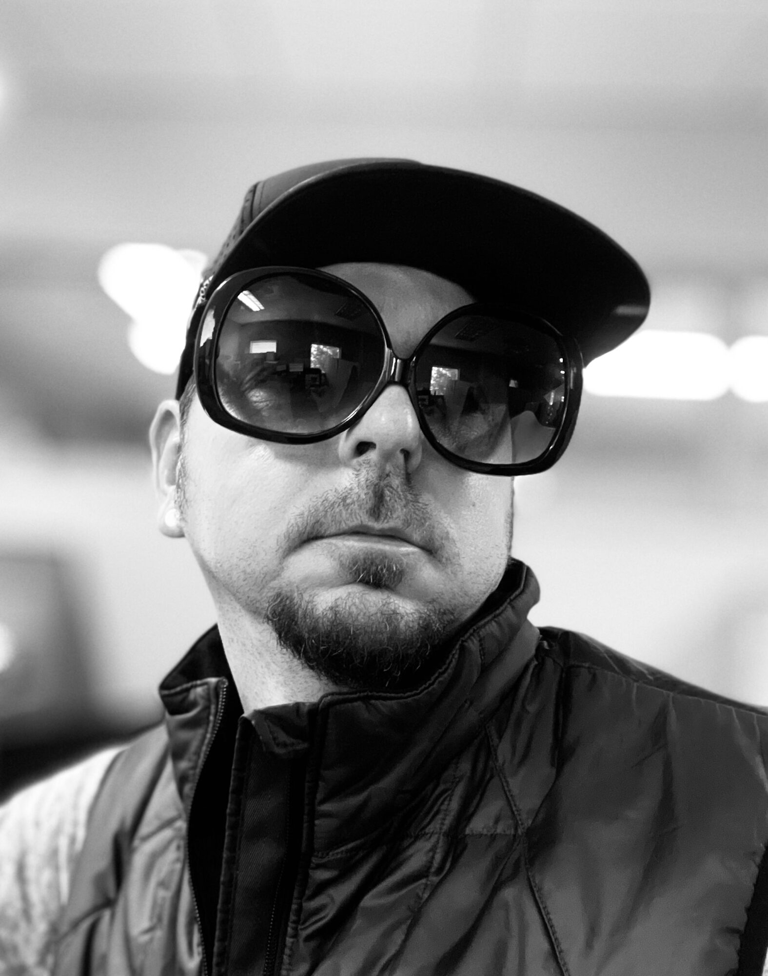 Man wearing large sunglasses, cap, and jacket, looking at the camera in black and white.