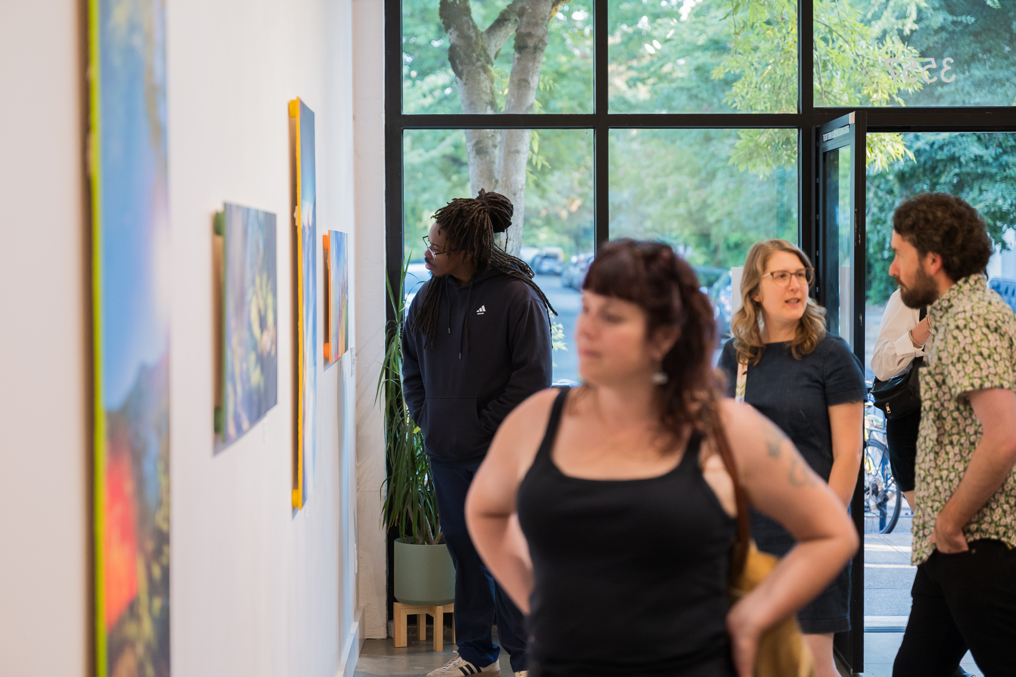 Group of people viewing artwork in gallery with large window and trees outside, some looking at paintings, others talking.