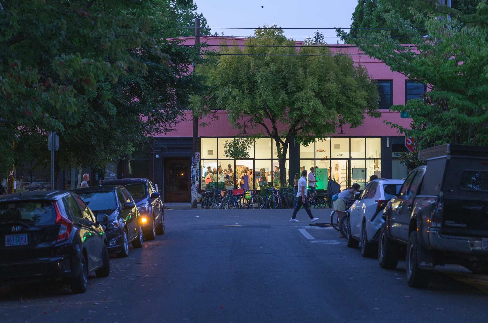 Building with large glass windows, trees in front, people walking, parked cars on both sides of the street.