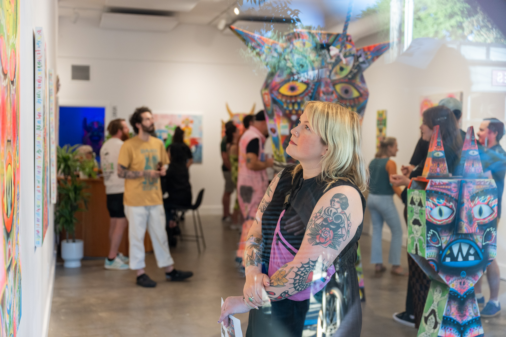 Woman with tattoos and colorful sleeves looking at artwork in an art gallery with people in the background.