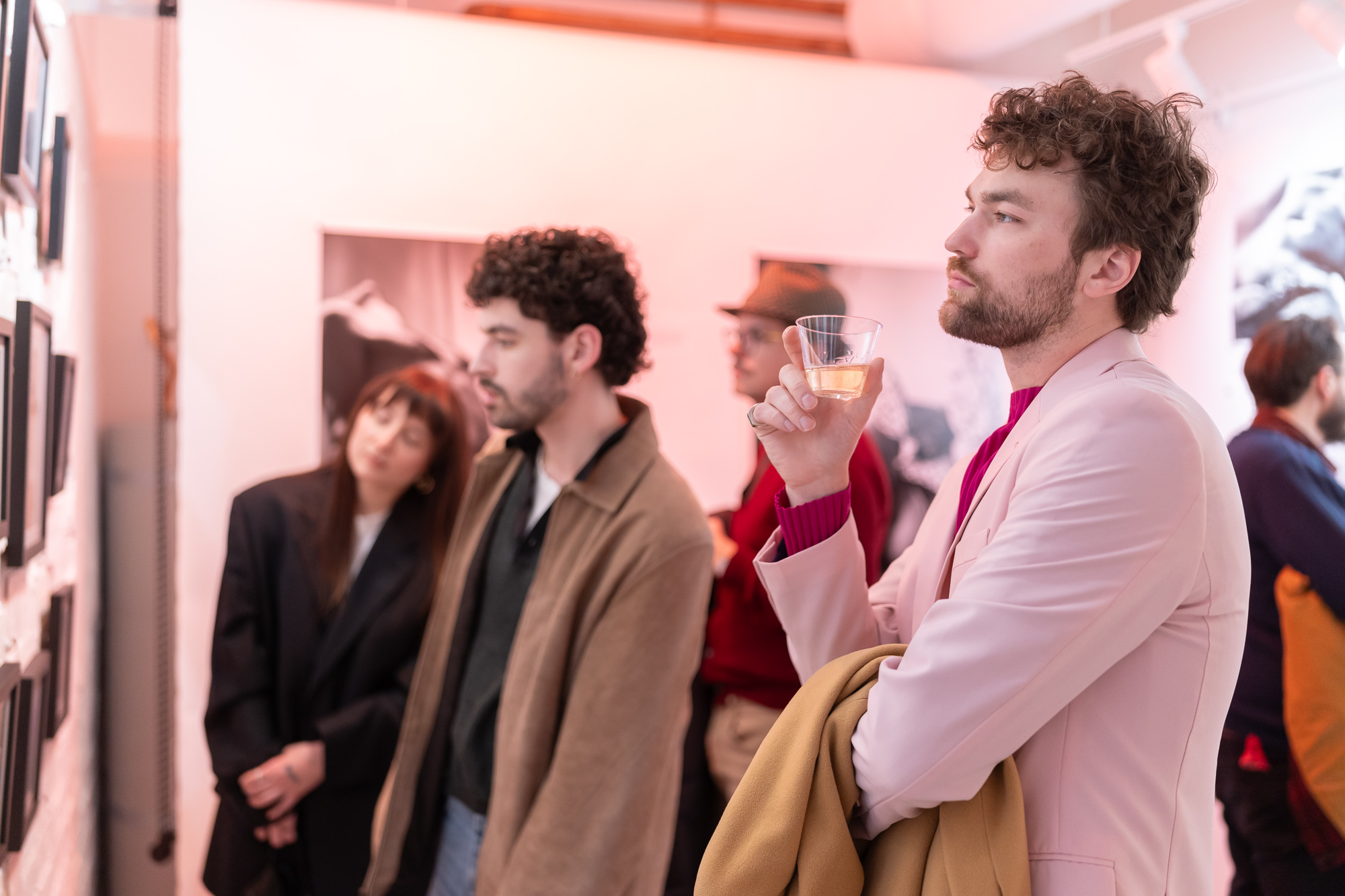 Man in pink blazer holding a drink, surrounded by people in an indoor setting.