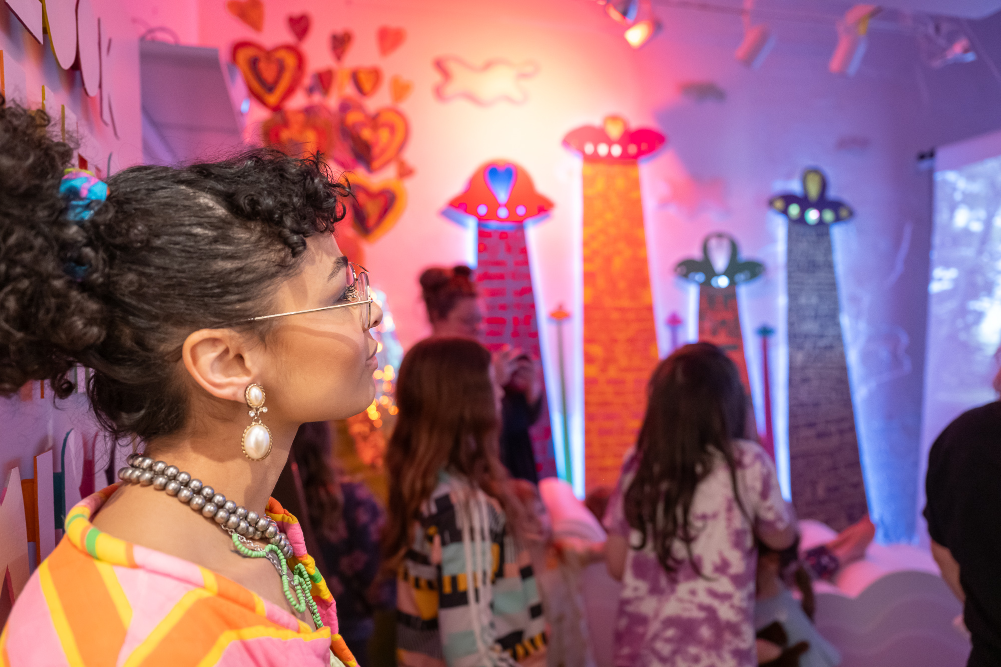 Group of people, mostly women, in a colorful room with heart and star decorations, some facing a screen or display.