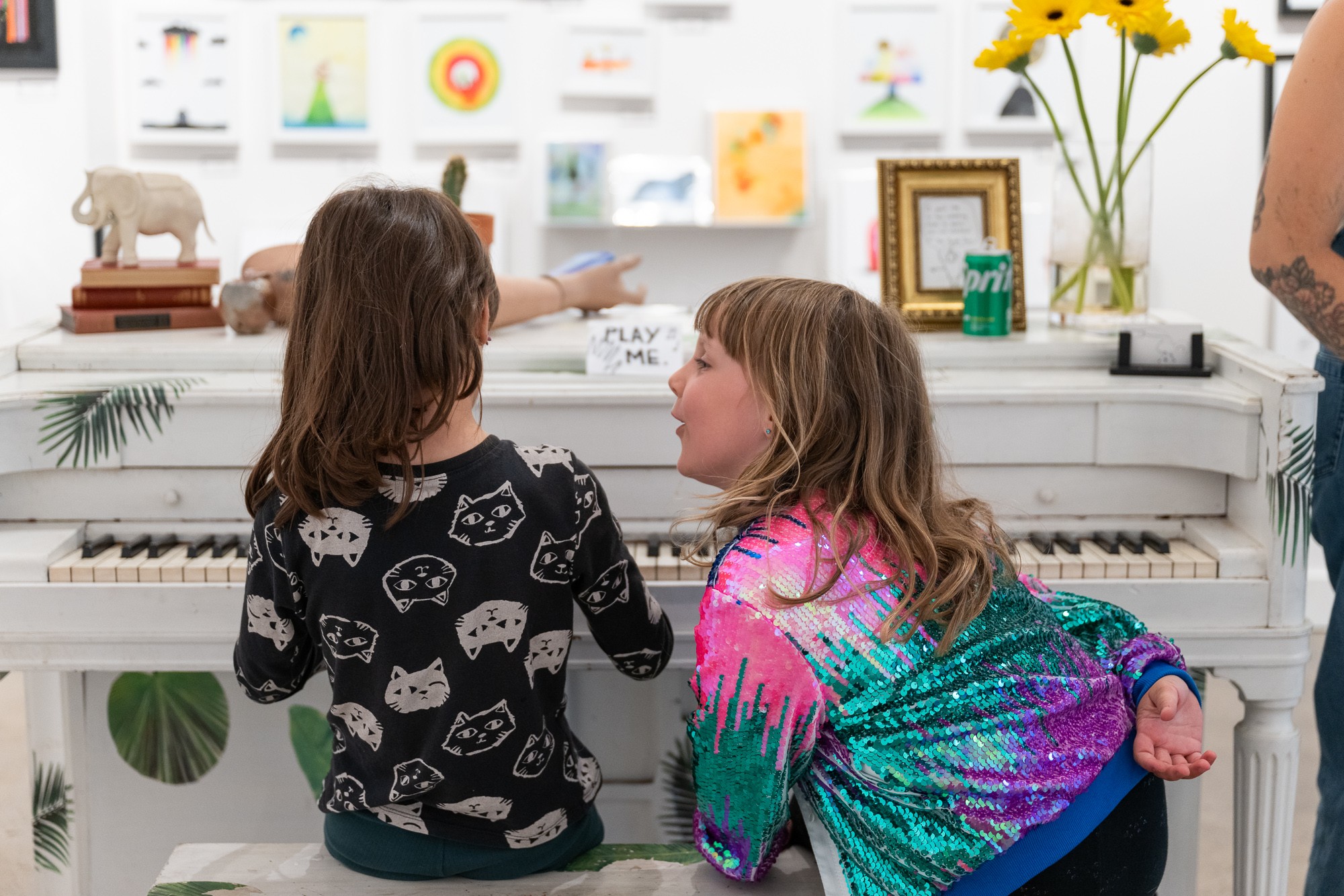 Two children sit at a white piano, facing each other, in a room with artwork and flowers on the wall.