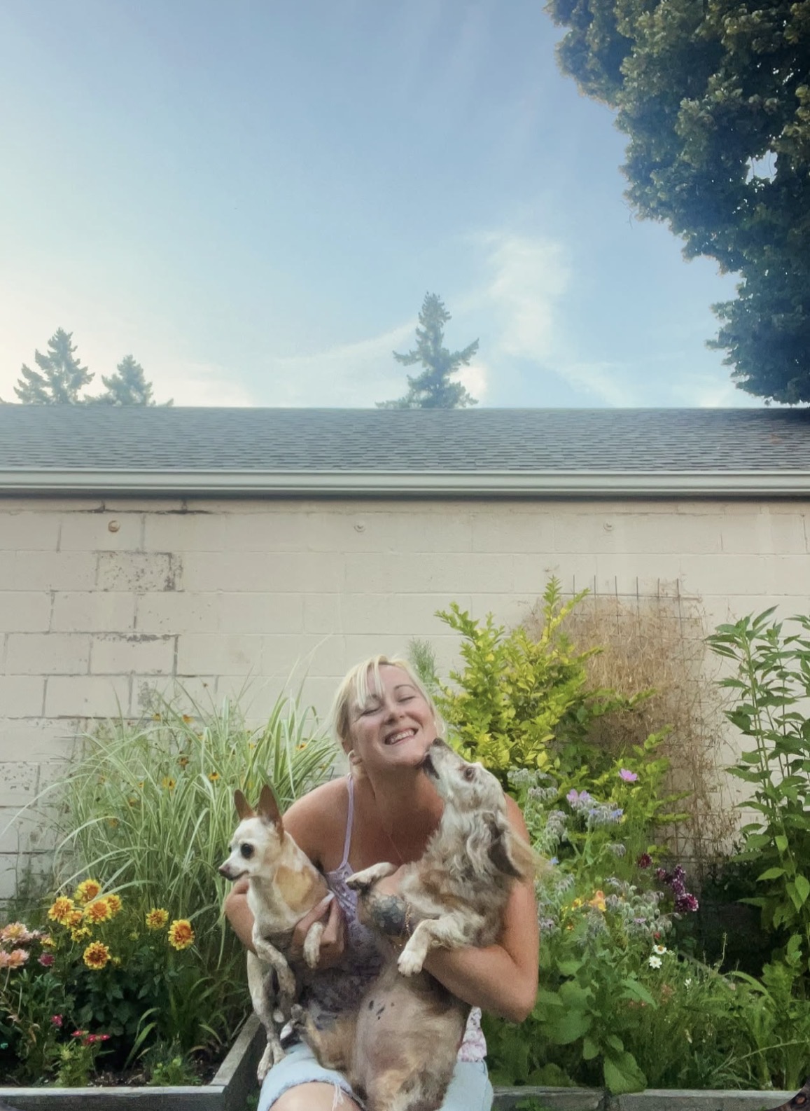 Person smiling and holding two dogs outdoors in a garden with flowers and greenery, sky visible above.