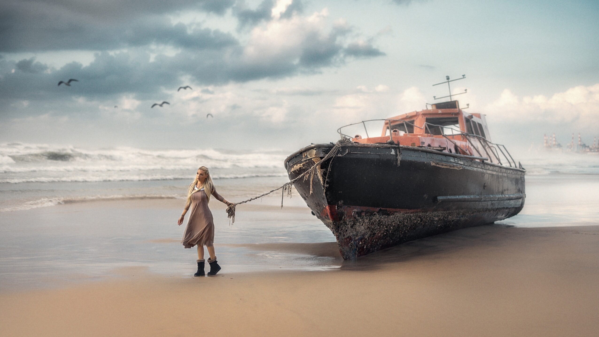 Child holding a stick near a boat on the beach, with clouds and ocean in the background.