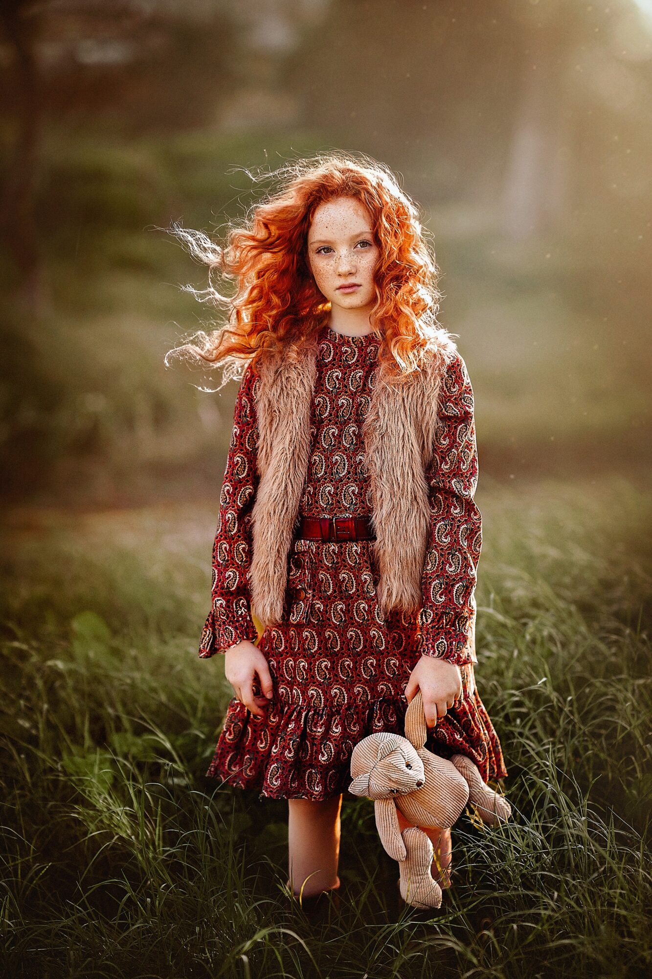 Young woman with curly red hair standing outdoors in a grassy field, holding a stuffed animal, wearing a patterned dress and vest.
