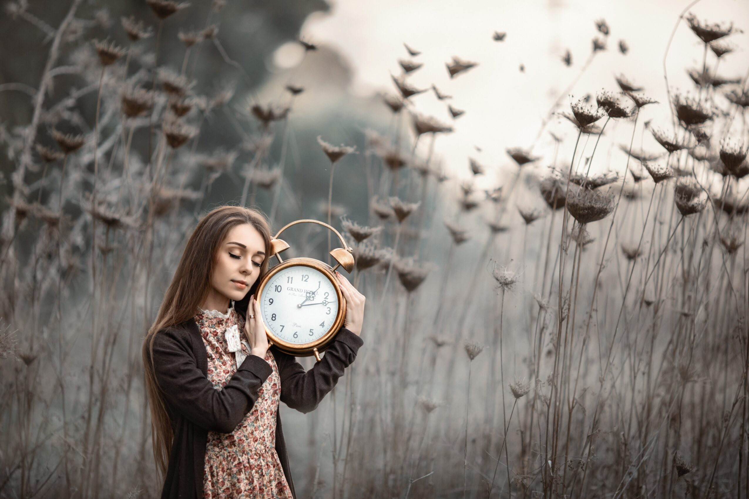 Young woman with long hair holding a clock outdoors in a field with tall plants and flying insects, overcast sky.