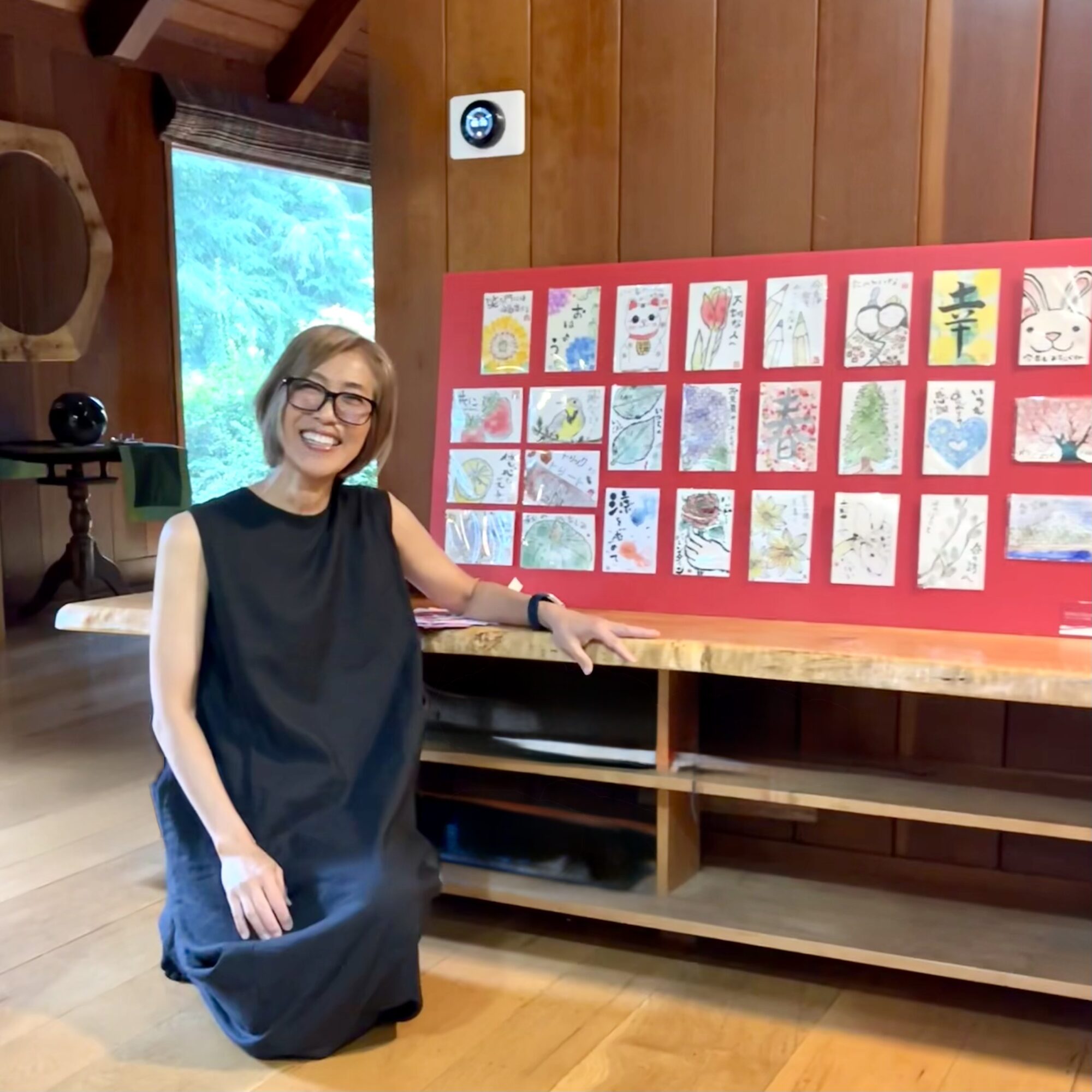 Woman kneeling next to a display board with colorful artwork, smiling, in a wooden room with a window and shelves.