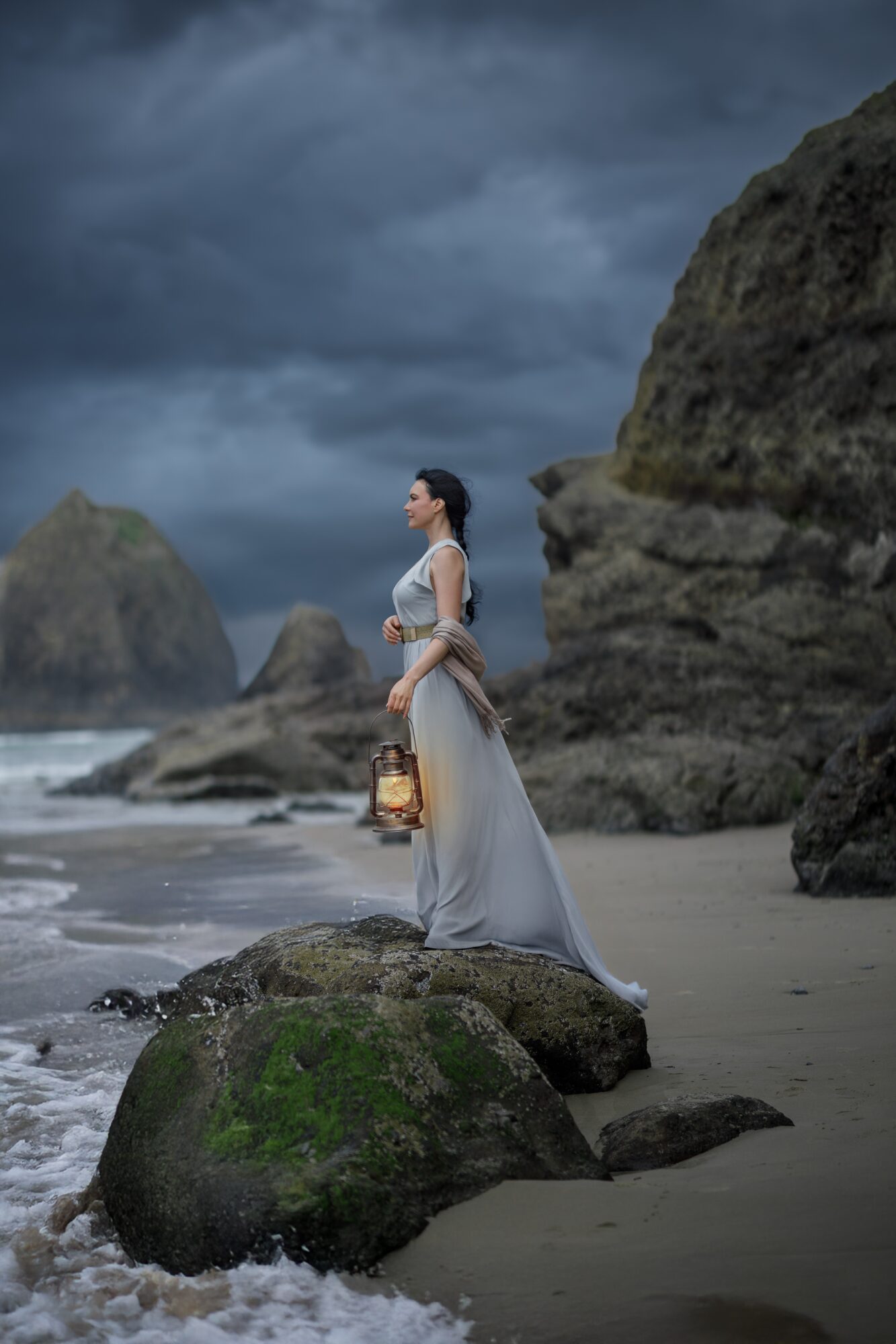 Woman in a long dress holding a lantern stands on rocks at the beach with large rocks and dark cloudy sky behind her.
