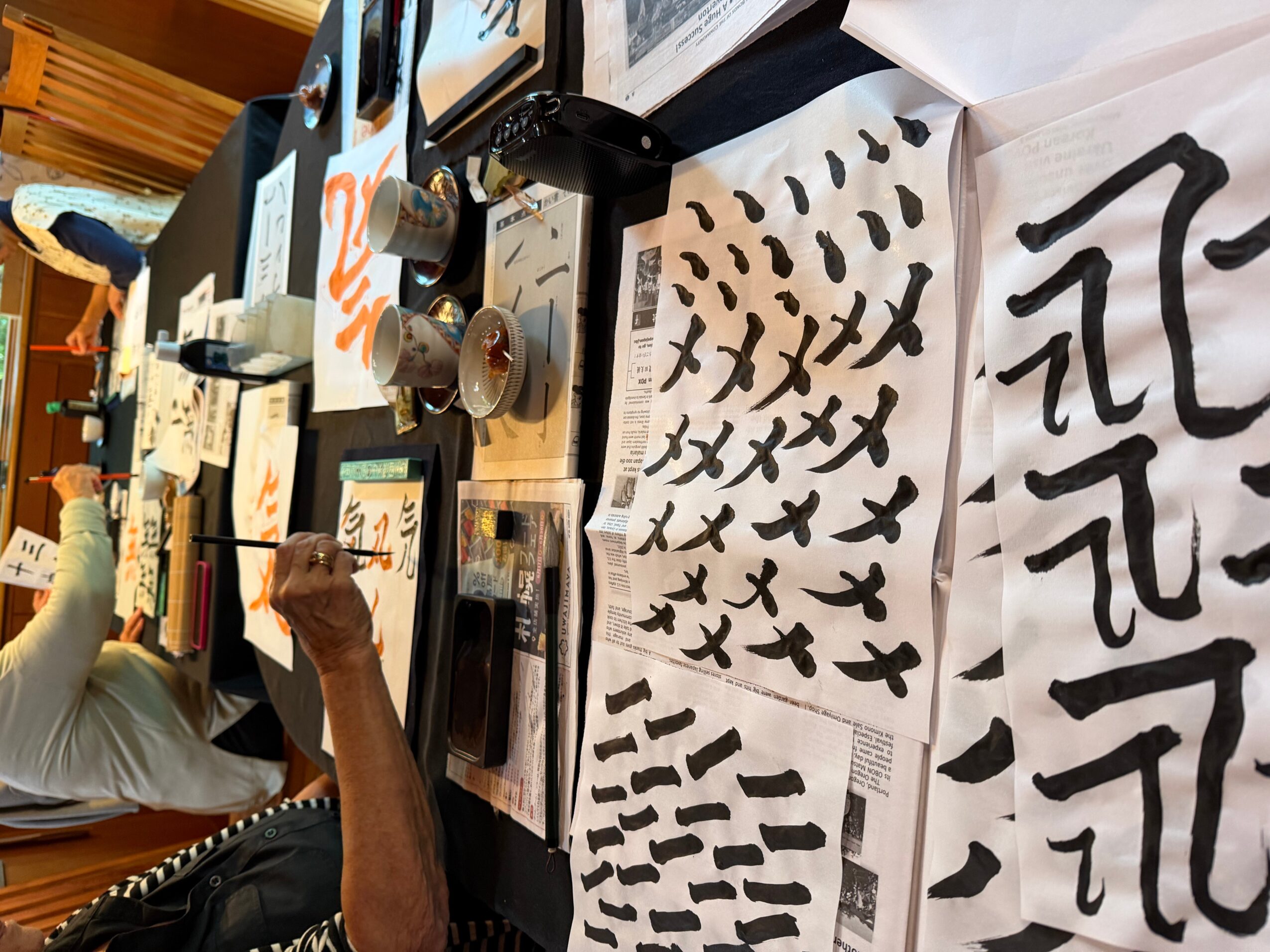 Person practicing calligraphy with black ink on paper, various brush strokes and characters visible, workspace with brushes and ink.