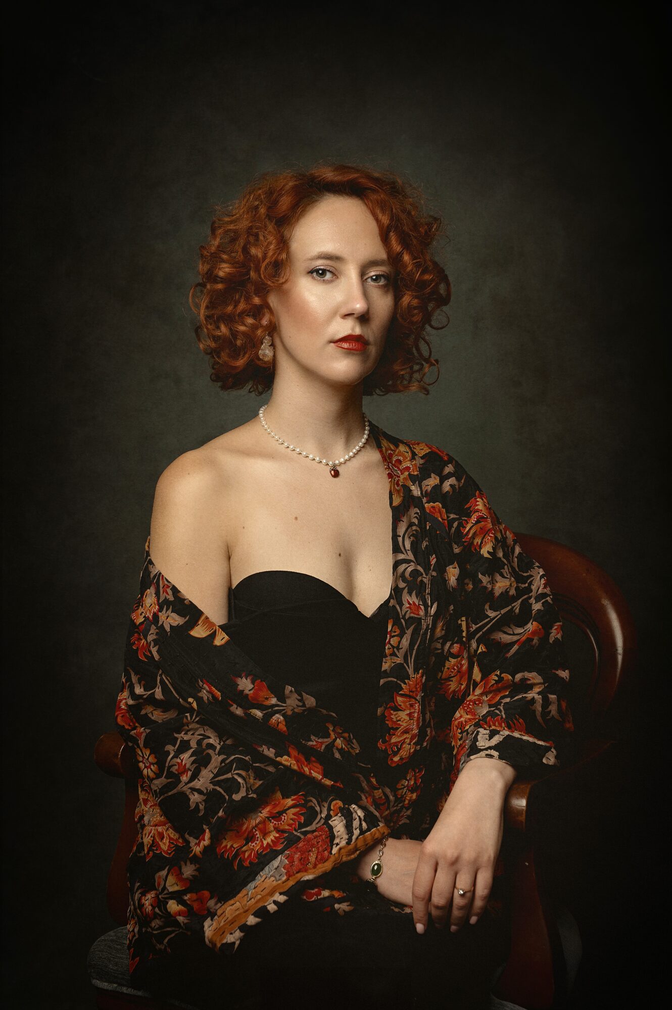 Portrait of a woman with curly red hair, wearing a black strapless top and a patterned shawl, sitting on a chair.