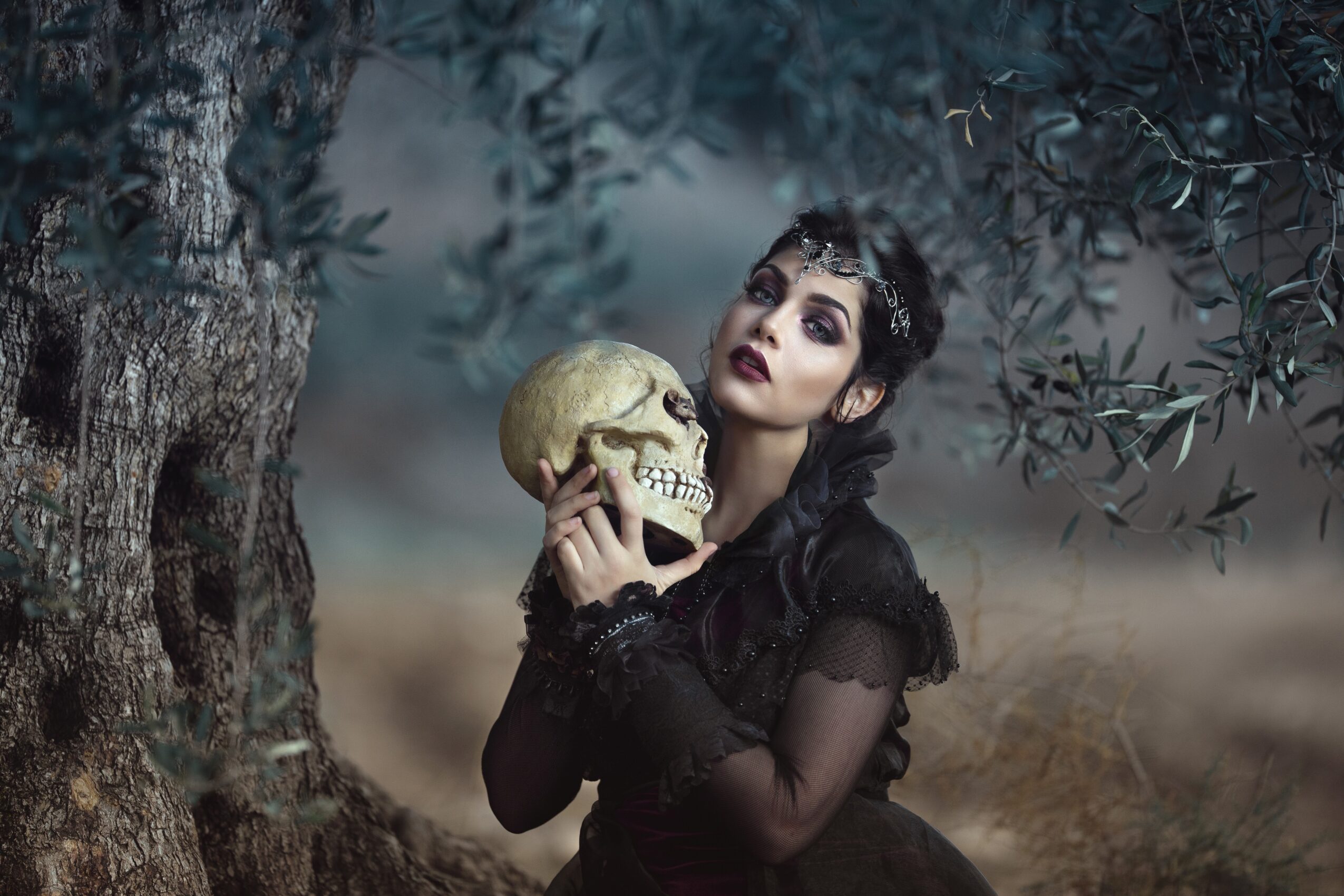 A woman with dark makeup and black clothing holds a skull near her face outdoors, with trees and cloudy sky in background.