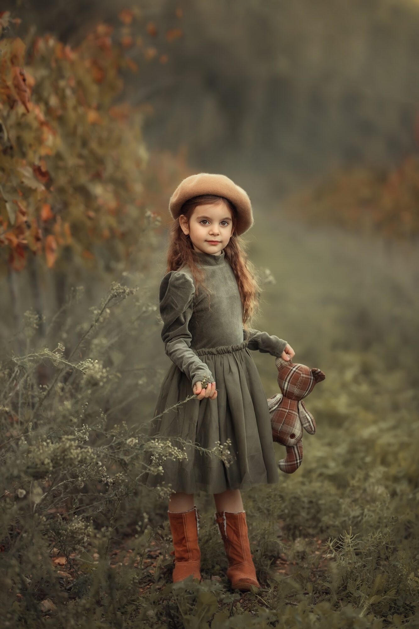 Young girl in a dress and hat holding a teddy bear outdoors in autumn scenery.