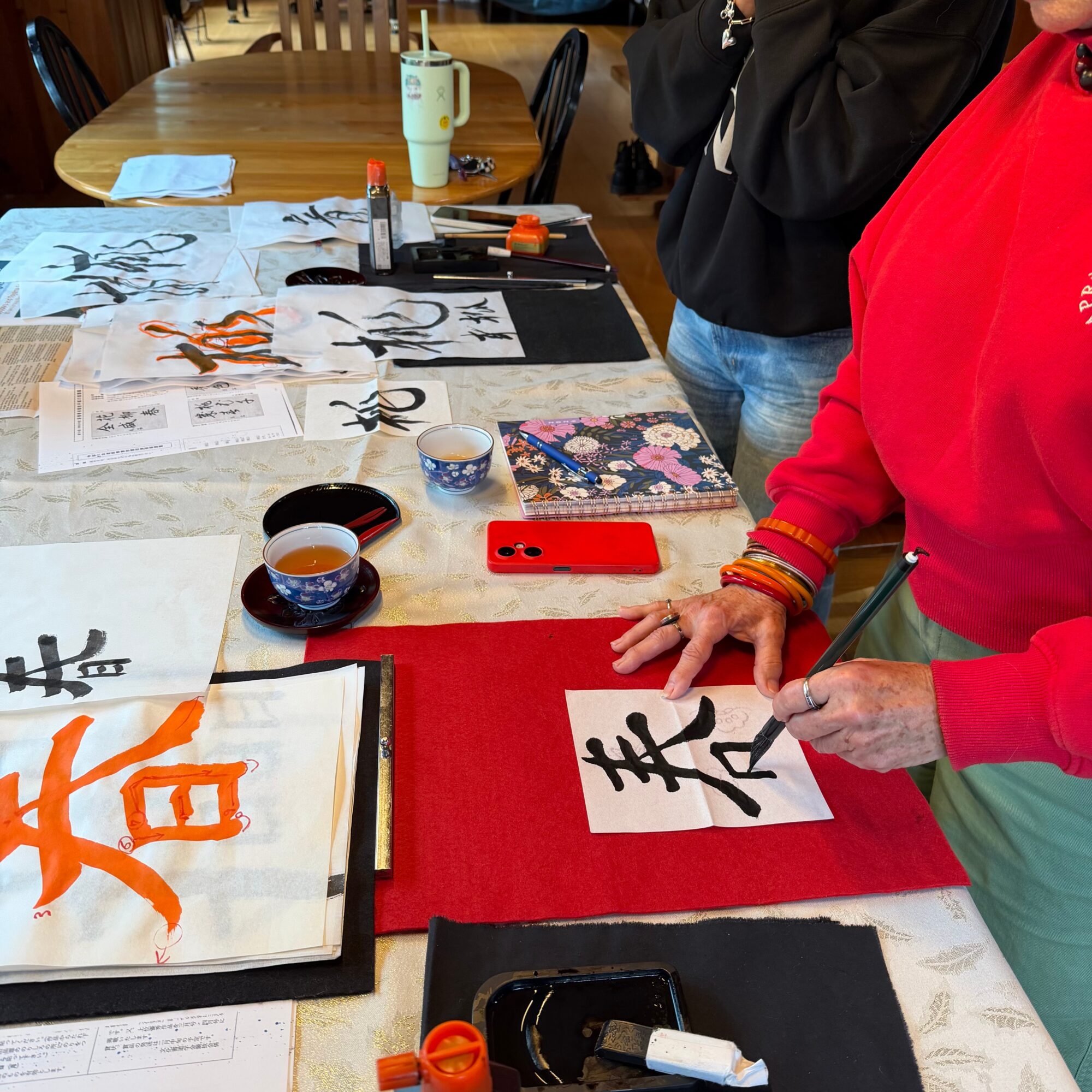 Person practicing calligraphy with brushes and ink on paper, surrounded by calligraphy materials on a table.