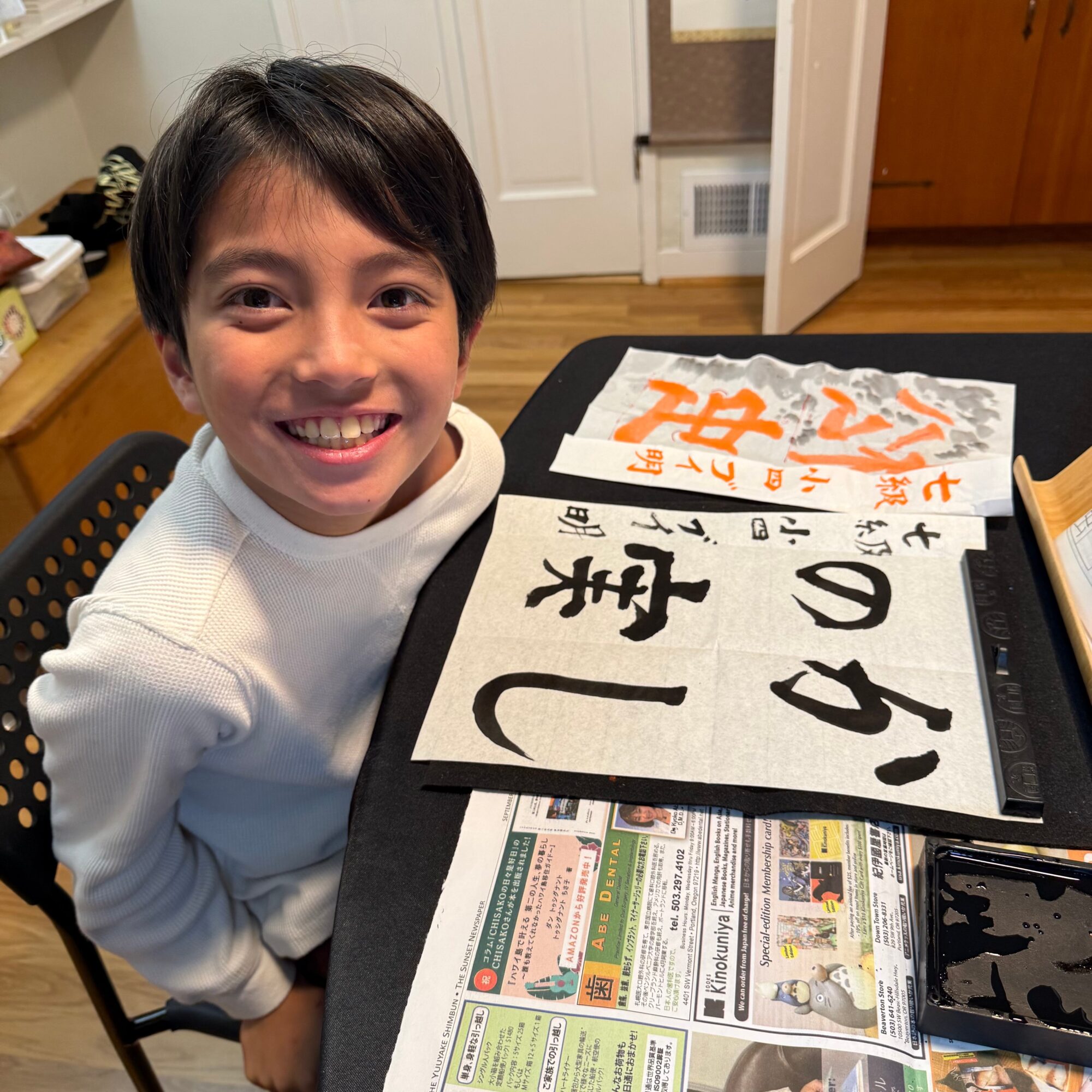 Smiling boy sitting at a table with calligraphy papers and newspaper, looking at the camera.