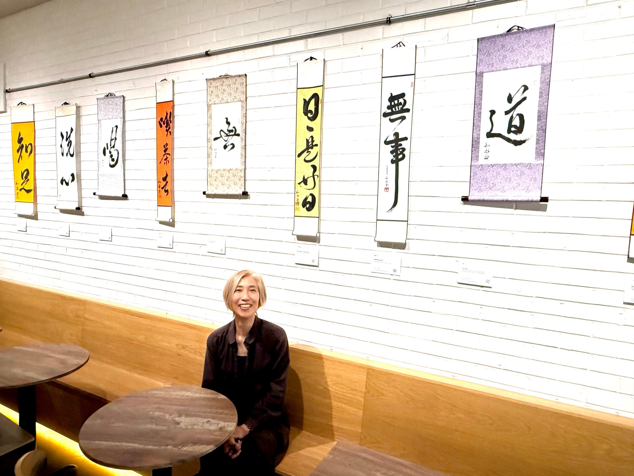 A smiling woman sits at a round table in a room with white brick walls and hanging scrolls with Japanese calligraphy.