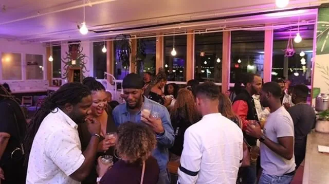 Group of people socializing indoors with large windows and colorful lighting, some holding drinks, in a lively setting.