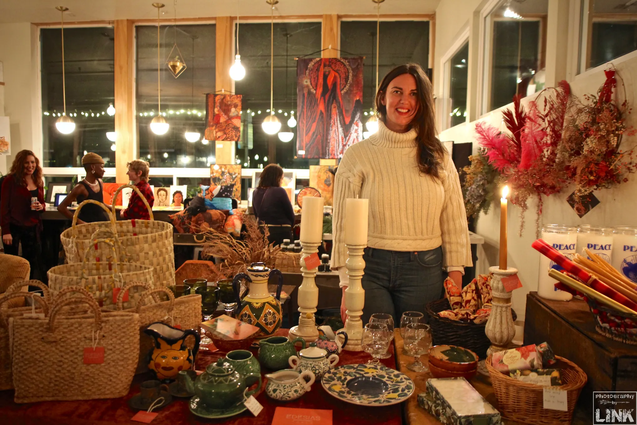 Woman standing behind a table with various decorative items in a lively indoor setting, with people seated in the background.