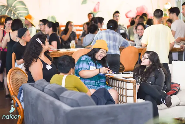Group of people socializing indoors, some sitting on chairs and couches, others standing, with a lively atmosphere.